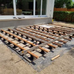 Budowa tarasu: widok na konstrukcję z drewnianych legarów, poziomicy i wkrętarki akumulatorowej na regulowanych wspornikach na czarnej geowłókninie, z kostką brukową w tle i oknami budynku