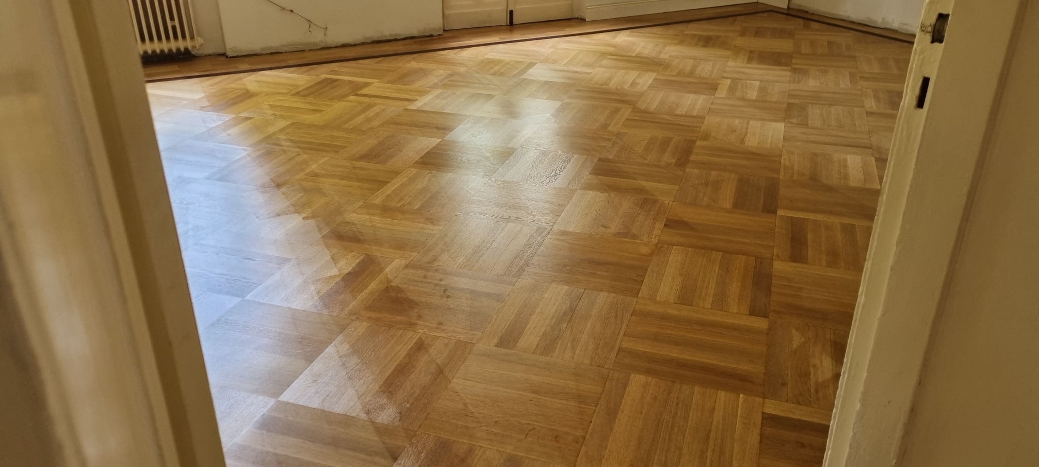 Parkiet ułożony w kwadraty, wykończony ciemną listwą przypodłogową, widok zza uchylonych drzwi, ślady niedokończonych prac na ścianie.