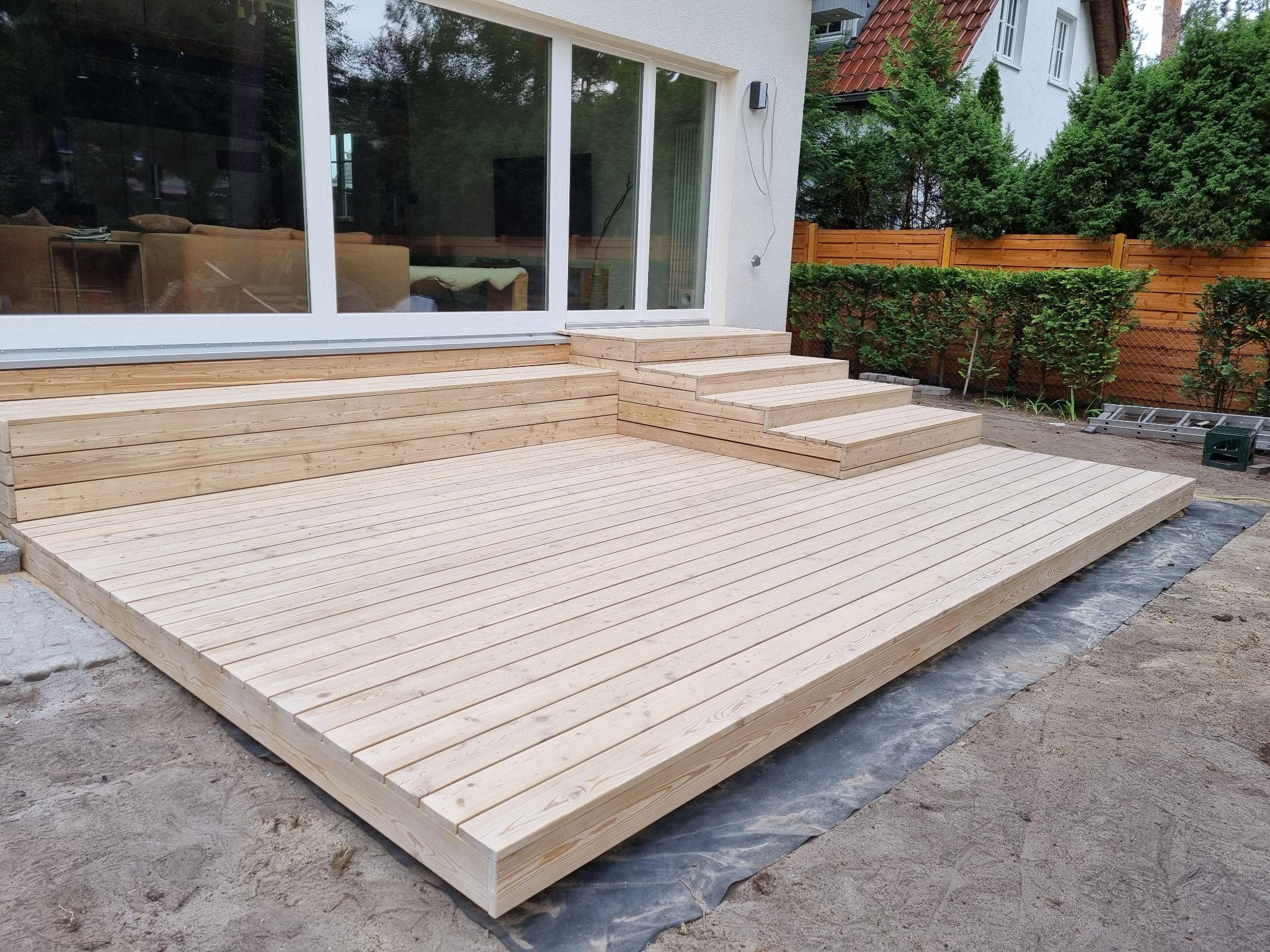 Nowo wybudowany drewniany taras z jasnych desek z niskimi schodami, przylegający do domu z dużymi przesuwnymi drzwiami tarasowymi, widoczne zabezpieczenie podłoża czarną folią.