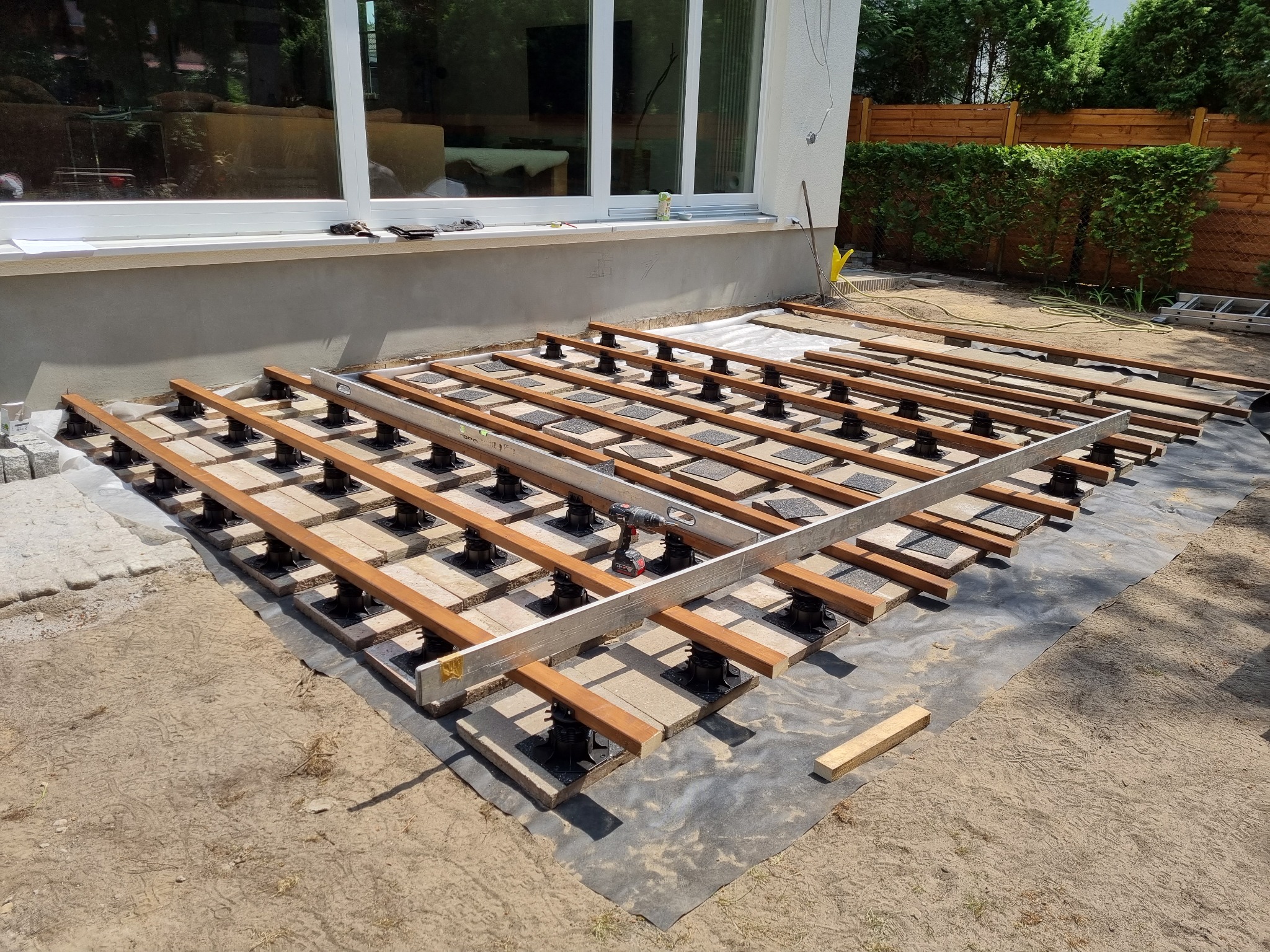 Budowa tarasu: widok na konstrukcję z drewnianych legarów, poziomicy i wkrętarki akumulatorowej na regulowanych wspornikach na czarnej geowłókninie, z kostką brukową w tle i oknami budynku