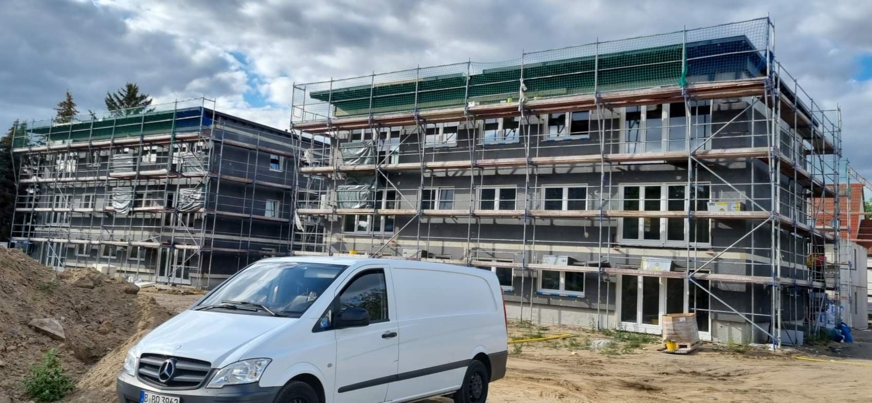 Dwa budynki wielorodzinne w trakcie prac wykończeniowych, elewacja w kolorze szarym, okna częściowo zasłonięte folią, rusztowania z siatką ochronną, na pierwszym planie biały samochód dostawczy.