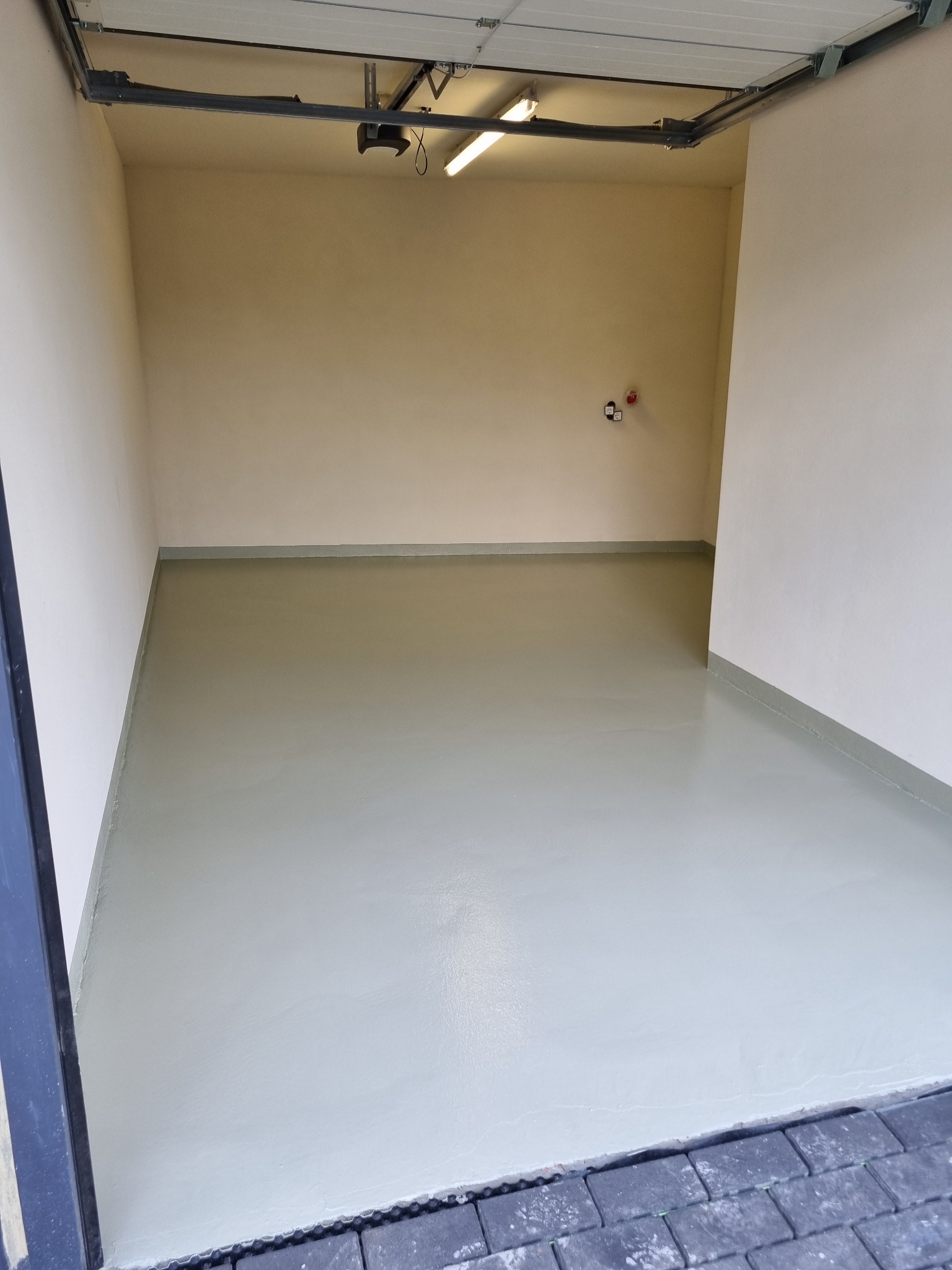 Pomalowana na szaro posadzka w pustym garażu z beżowymi ścianami i widocznym mechanizmem bramy garażowej pod sufitem.