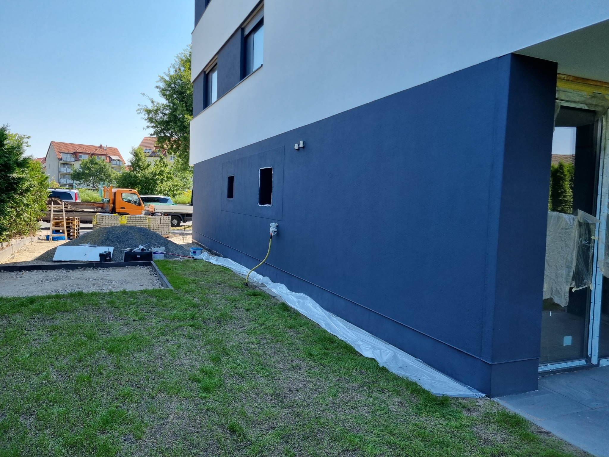 Świeżo pomalowana, granatowa elewacja budynku z widocznymi otworami okiennymi przed montażem okien, zabezpieczona folią malarską przy trawniku. Na trawniku widoczne ślady po pracach ziemnych.