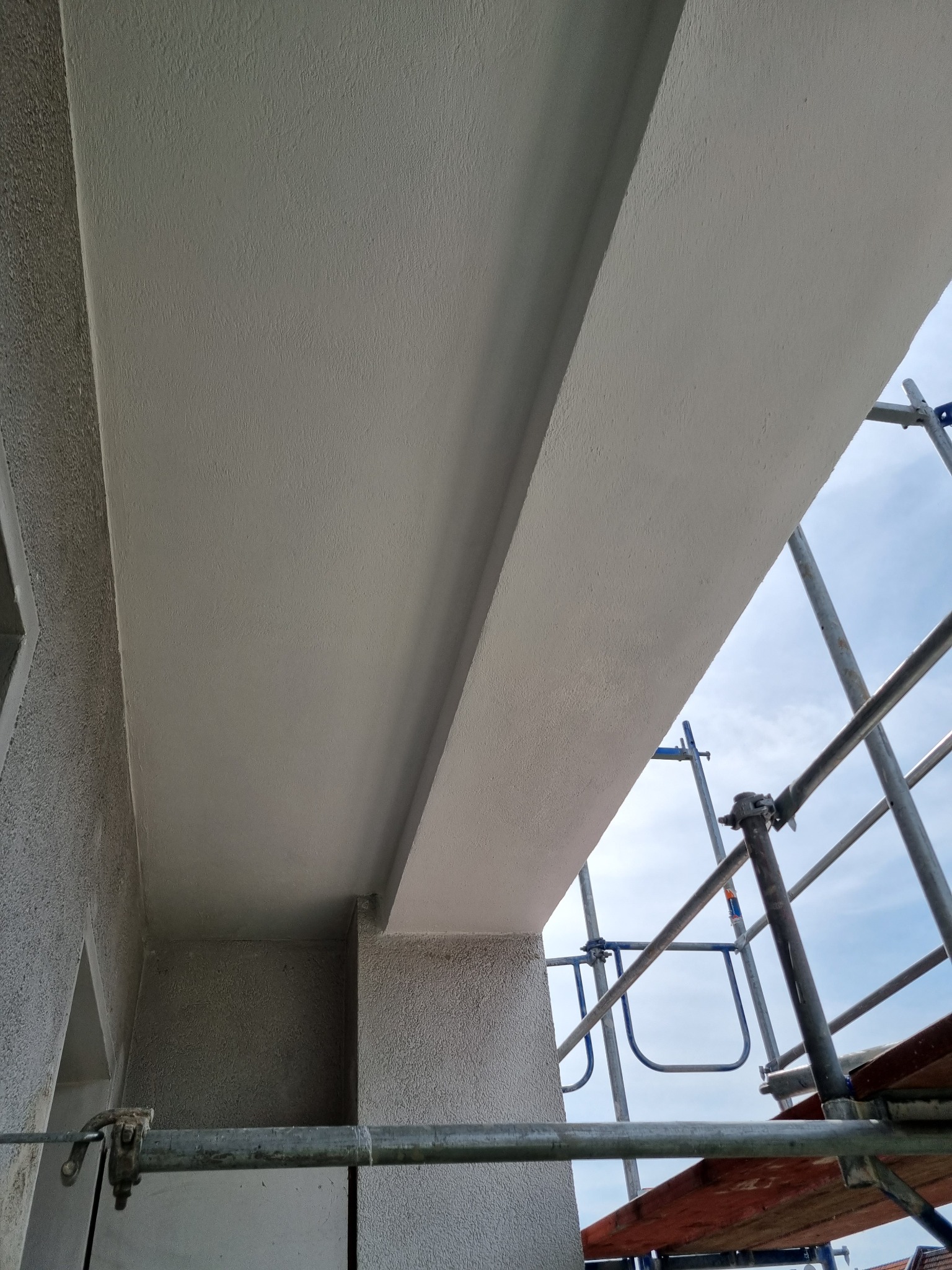 Świeżo otynkowany sufit i fragment ściany z rusztowaniem w tle, widoczne nierówności faktury tynku.