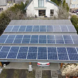 Widok z góry na instalację paneli słonecznych na dachu wiaty garażowej, z zaparkowanym pod nią srebrnym samochodem i stosem drewna opałowego obok.