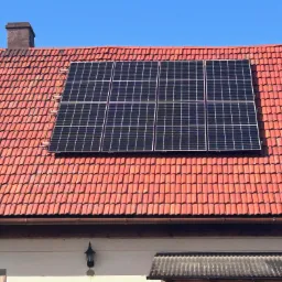 Panel słoneczny zamontowany na dachu z czerwonej dachówki, widok z dołu, na pierwszym planie fragment elewacji budynku.
