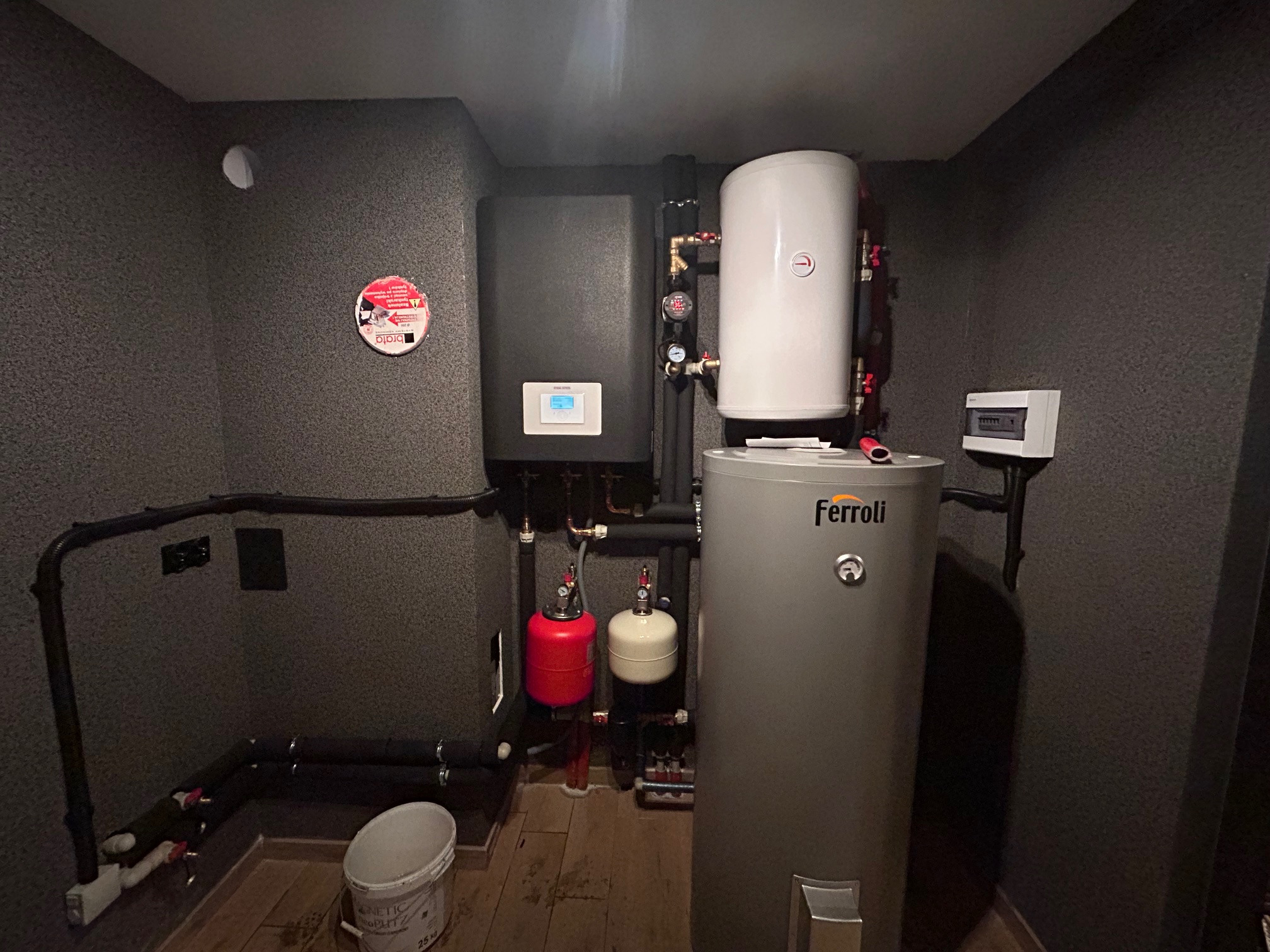 Zestaw urządzeń grzewczych w kotłowni: czarny piec z wyświetlaczem, biały i szary zbiornik, instalacja rur, małe zbiorniki kompensacyjne, kubeł na podłodze.