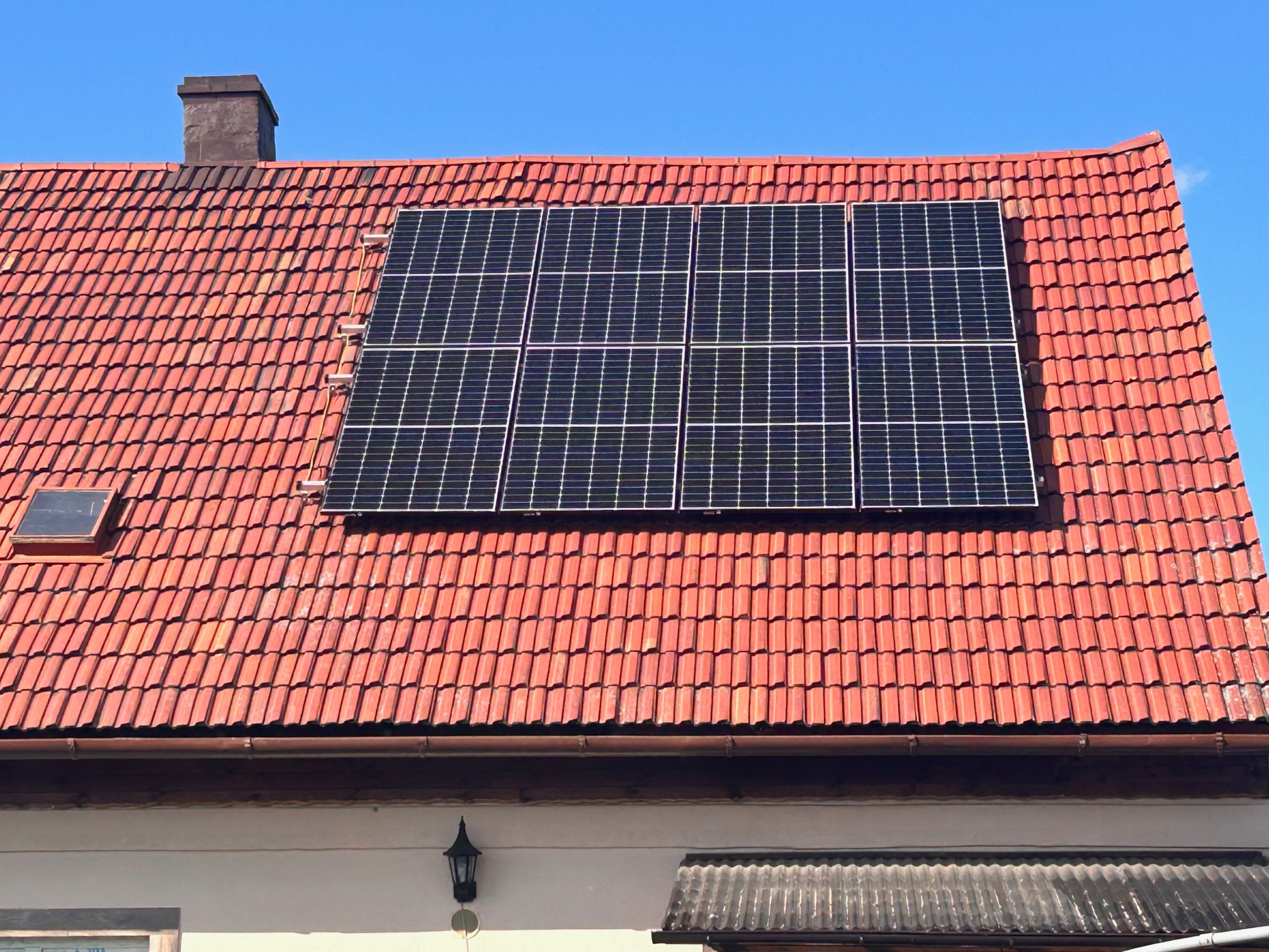 Panel słoneczny zamontowany na dachu z czerwonej dachówki, widok z dołu, na pierwszym planie fragment elewacji budynku.