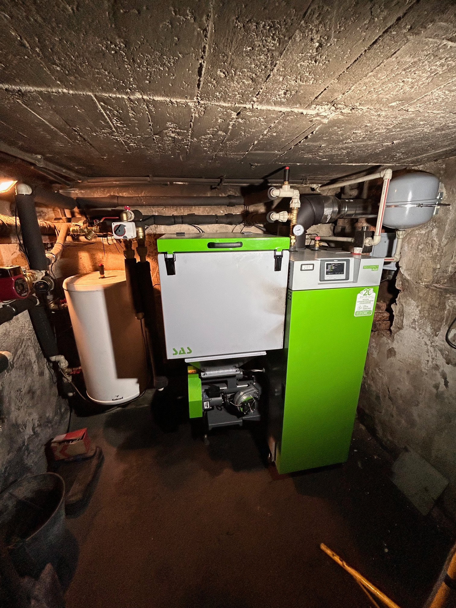 Nowoczesny piec CO z podajnikiem, w kolorach szaro-zielonych, zainstalowany w piwnicy o surowych ścianach i suficie z widocznymi instalacjami.