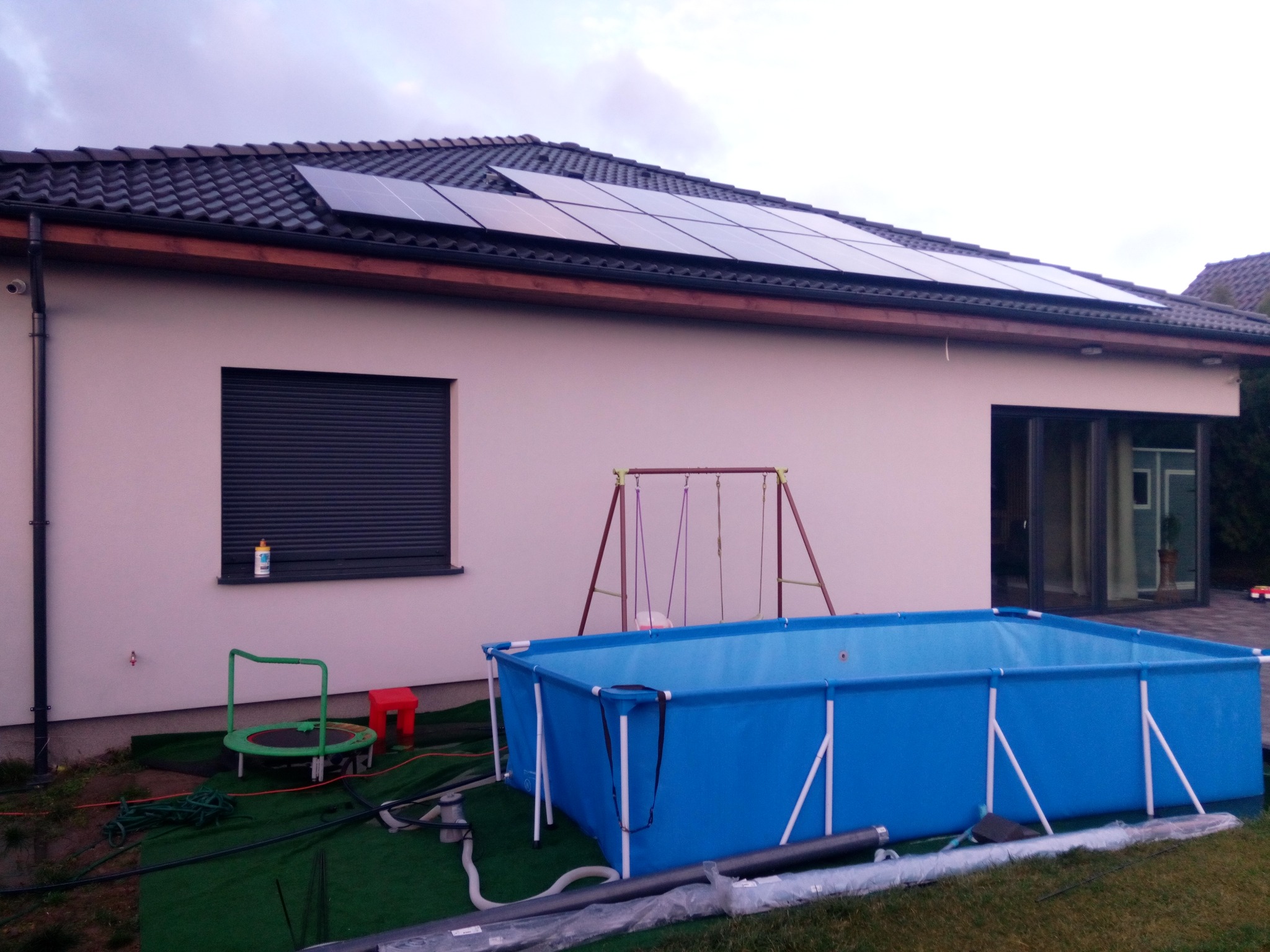 Dom jednorodzinny z panelami słonecznymi na dachu, basenem ogrodowym, trampoliną i huśtawką na podwórku, widok z boku.