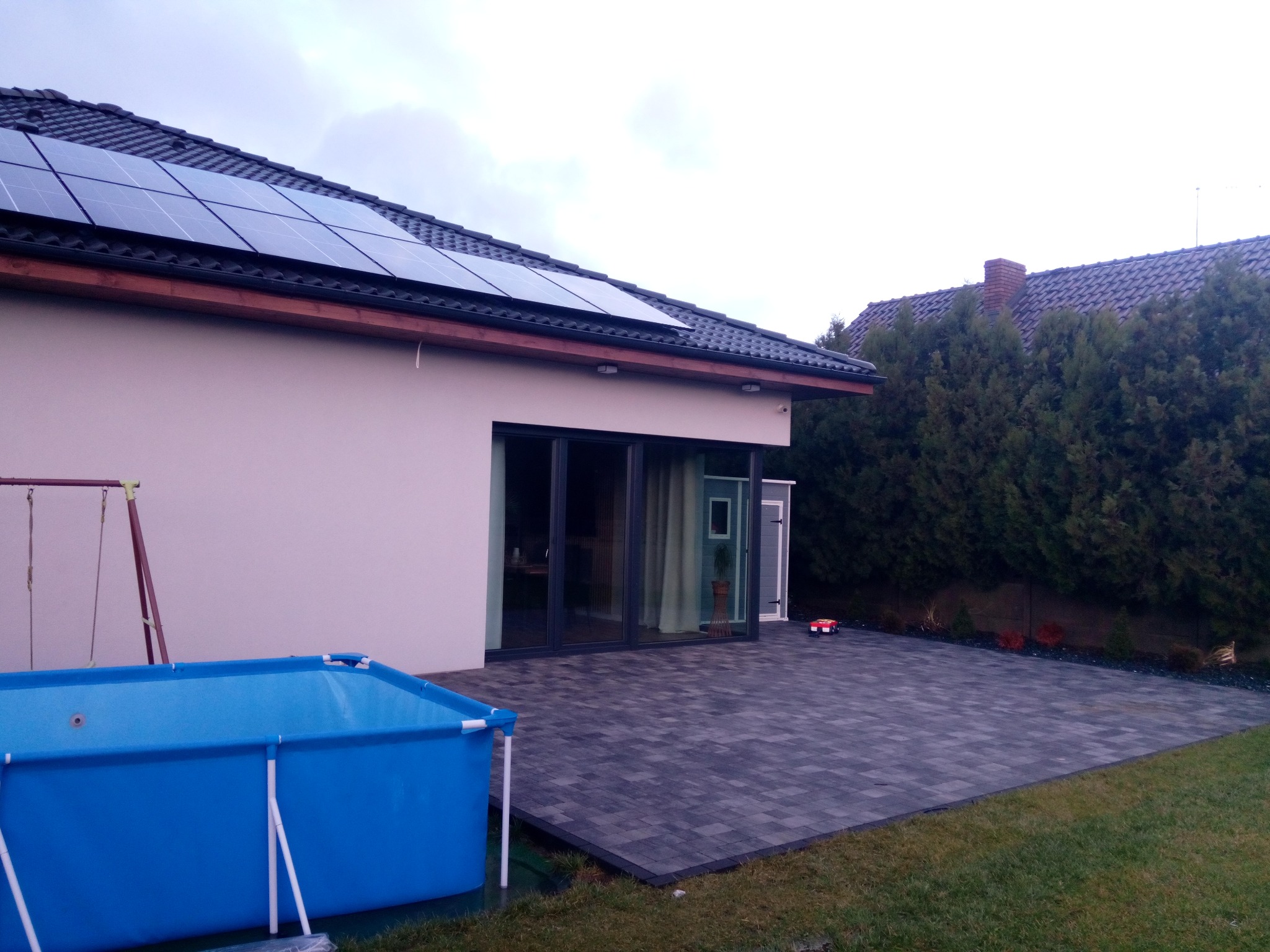 Nowoczesny dom jednorodzinny z panelami słonecznymi na dachu, tarasem z szarej kostki brukowej i basenem ogrodowym w kolorze niebieskim. W tle widoczny ogród z tujami i mała zabawka - czerwony...