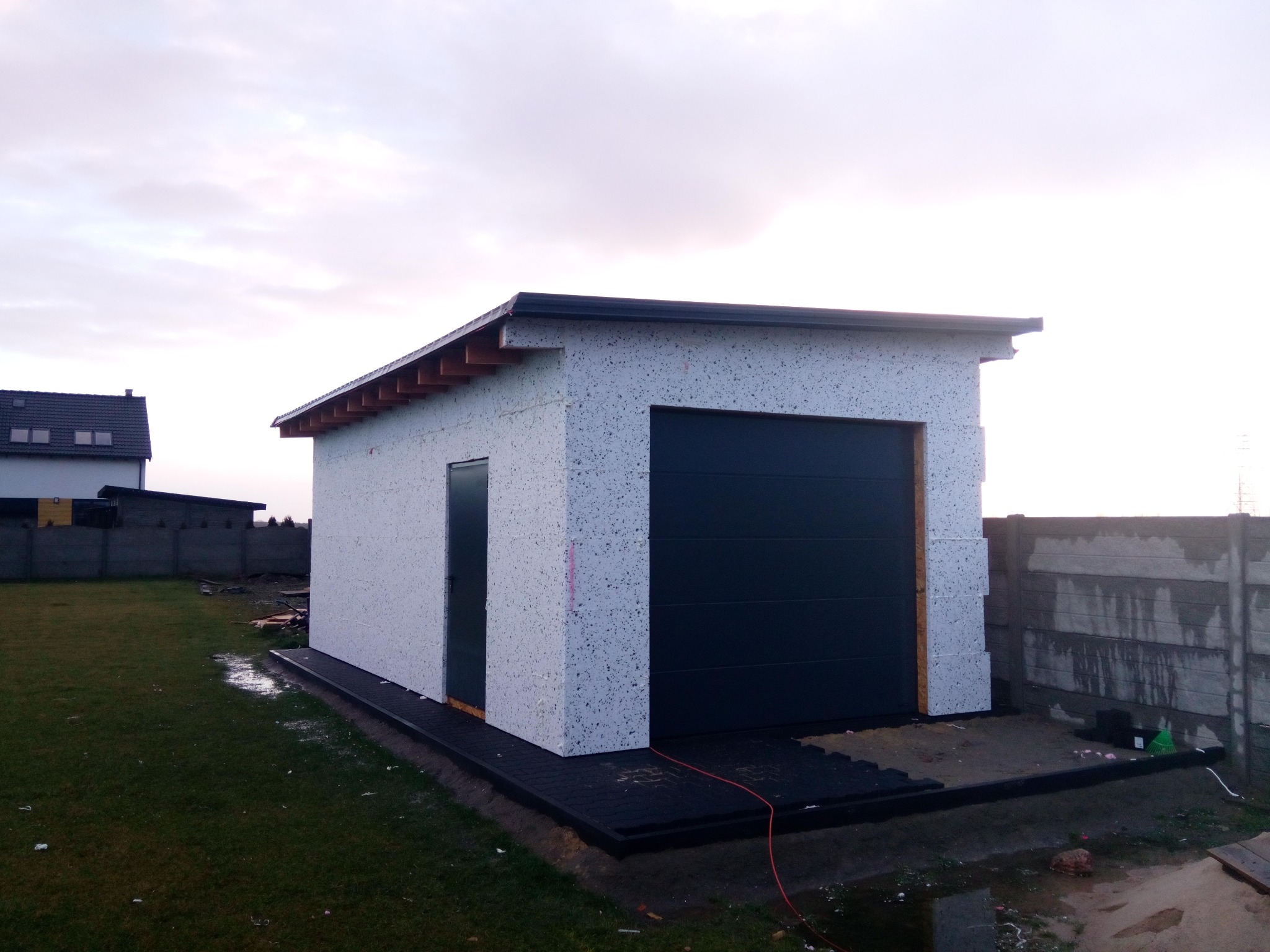 Niewielki, parterowy budynek gospodarczy z płaskim dachem, o ścianach z białego bloczka styropianowego i z czarną bramą garażową segmentową, na etapie budowy, z widocznymi elementami...