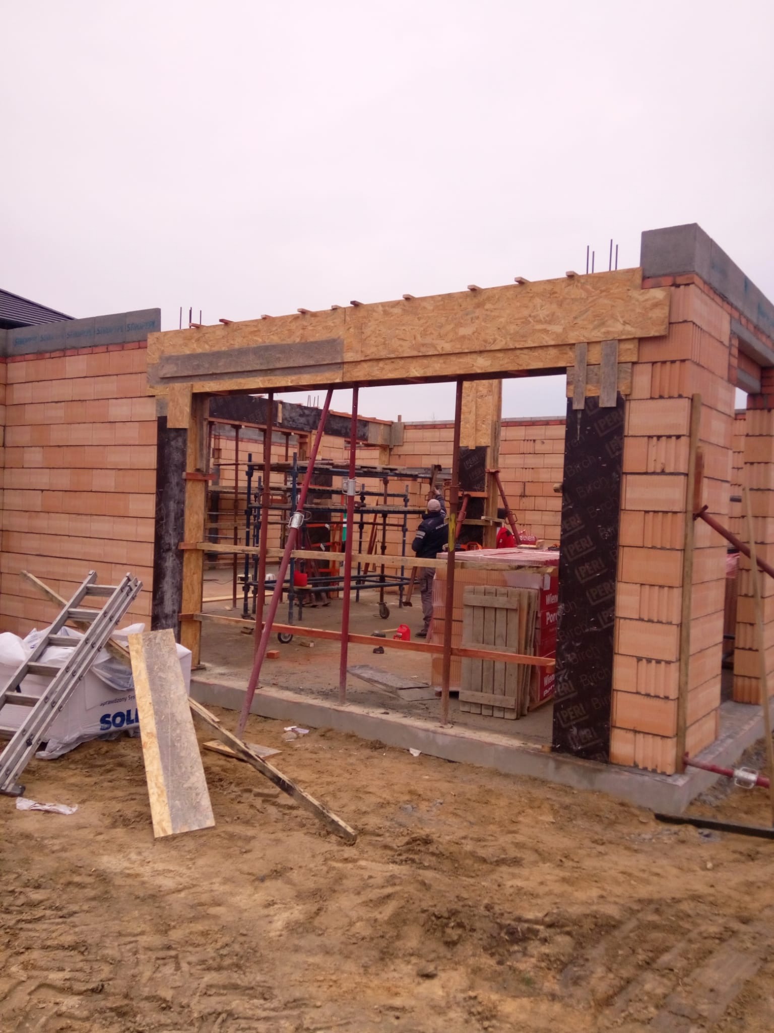 Wznoszenie ścian budynku z czerwonej cegły, widoczne rusztowania wewnątrz, drewniane szalunki nad otworem okiennym, drabina oparta o ścianę, ziemiste podłoże.