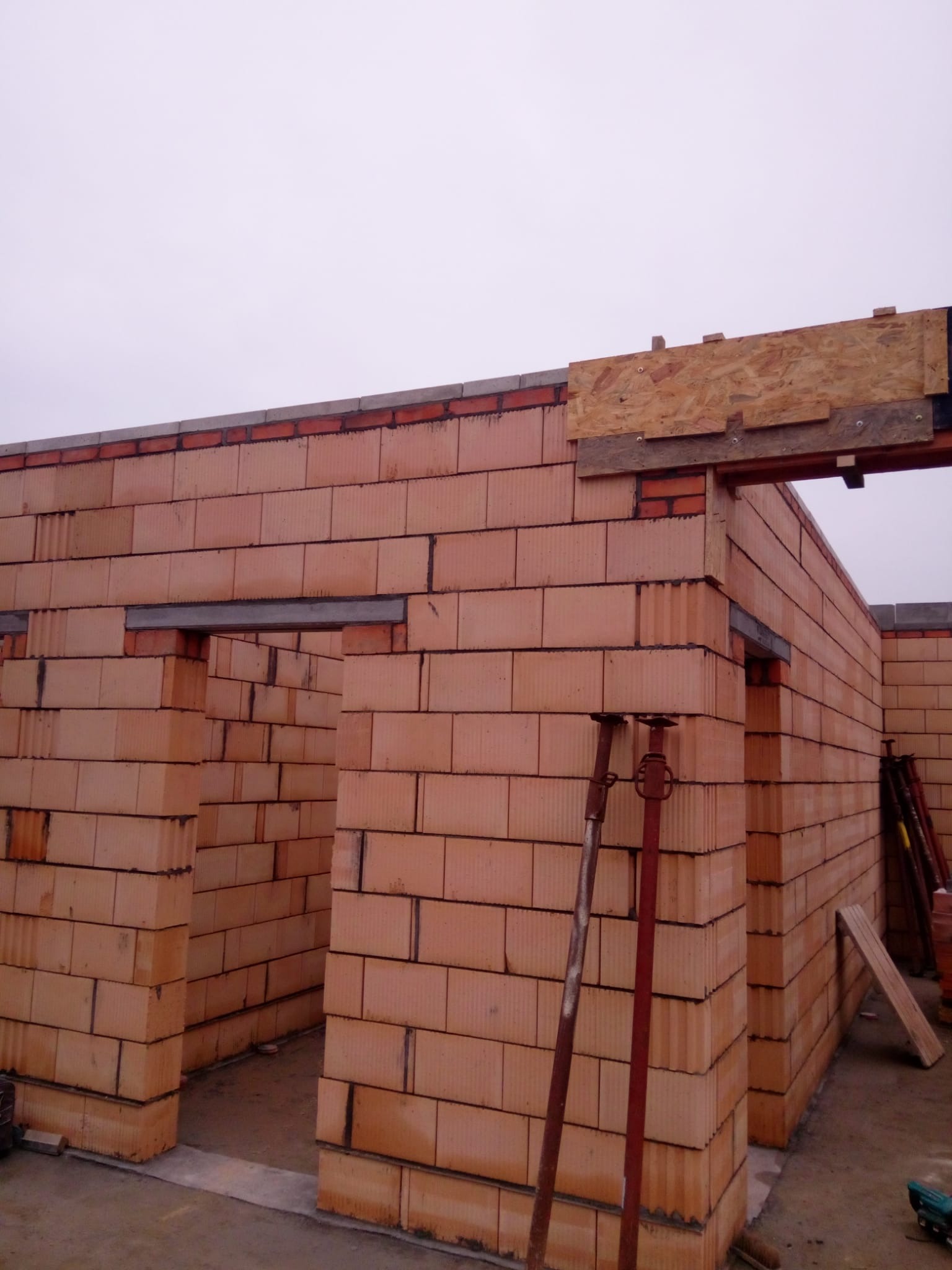 Wznoszone ściany budynku z pustaków ceramicznych, widoczne stalowe nadproża i podpory budowlane, szalunek z płyty OSB nad otworem okiennym.