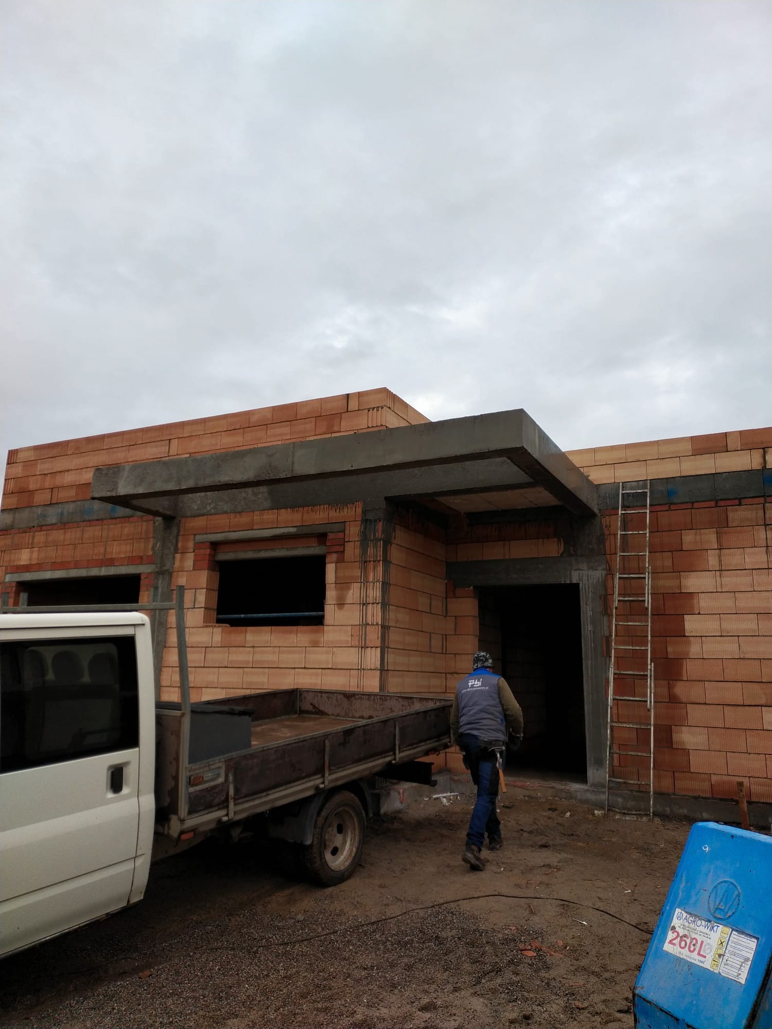 Budowa domu w trakcie wznoszenia ścian z czerwonej cegły, widoczny betonowy strop nad oknami i drzwiami, drabina oparta o ścianę, pracownik w kamizelce z logiem firmy oraz samochód dostawczy...