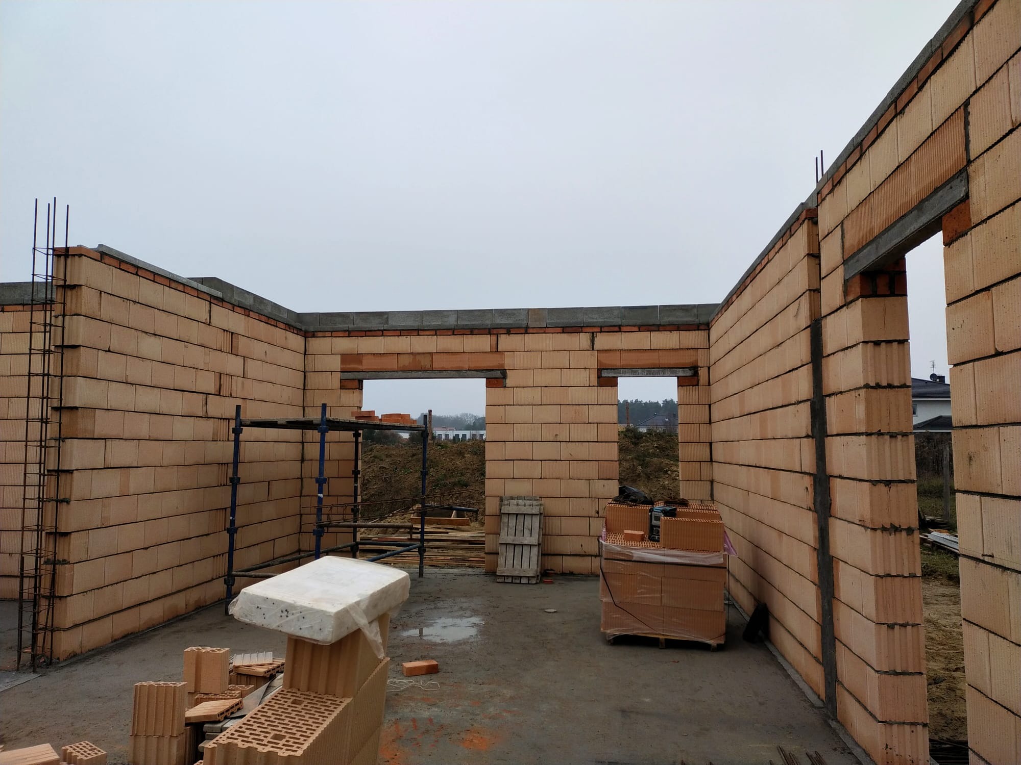 Wznoszone ściany domu z pustaków ceramicznych z widocznymi otworami okiennymi i zbrojeniami, na placu budowy w pochmurny dzień.