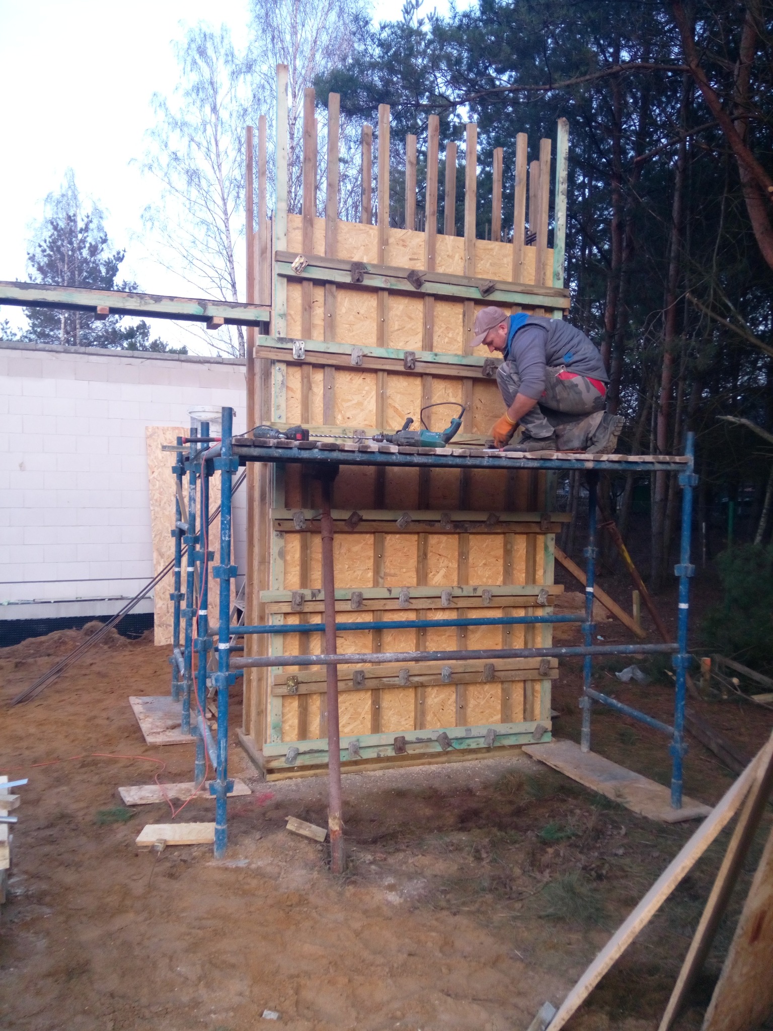 Pracownik na rusztowaniu, mocujący elementy szalunku z płyty OSB, przygotowanego do wylania betonu, widoczne narzędzia i elementy konstrukcyjne.