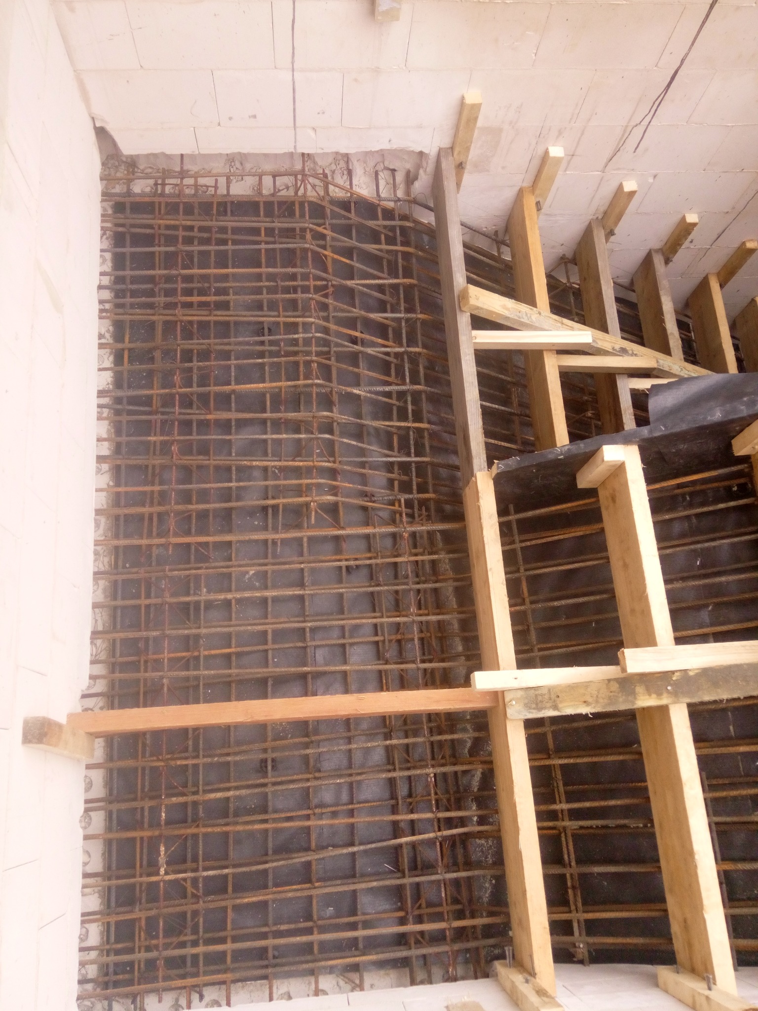 Siatka zbrojeniowa ułożona na folii w wykopie fundamentowym, widoczne drewniane szalunki i białe bloczki fundamentowe.