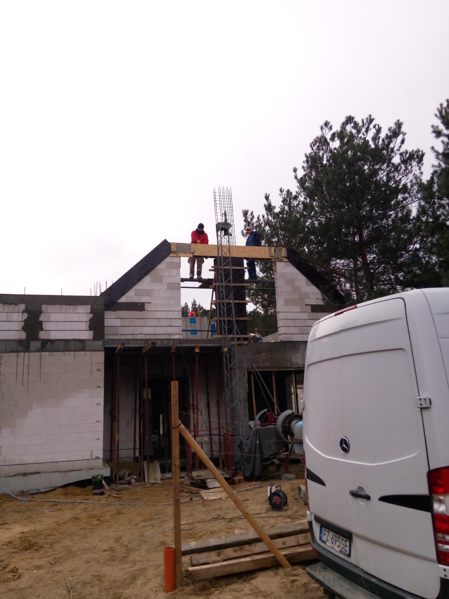 Wznoszenie ścian budynku z bloczków betonowych, widoczne rusztowanie, zbrojenie, oraz pracownicy na górze konstrukcji, w tle biały samochód dostawczy.