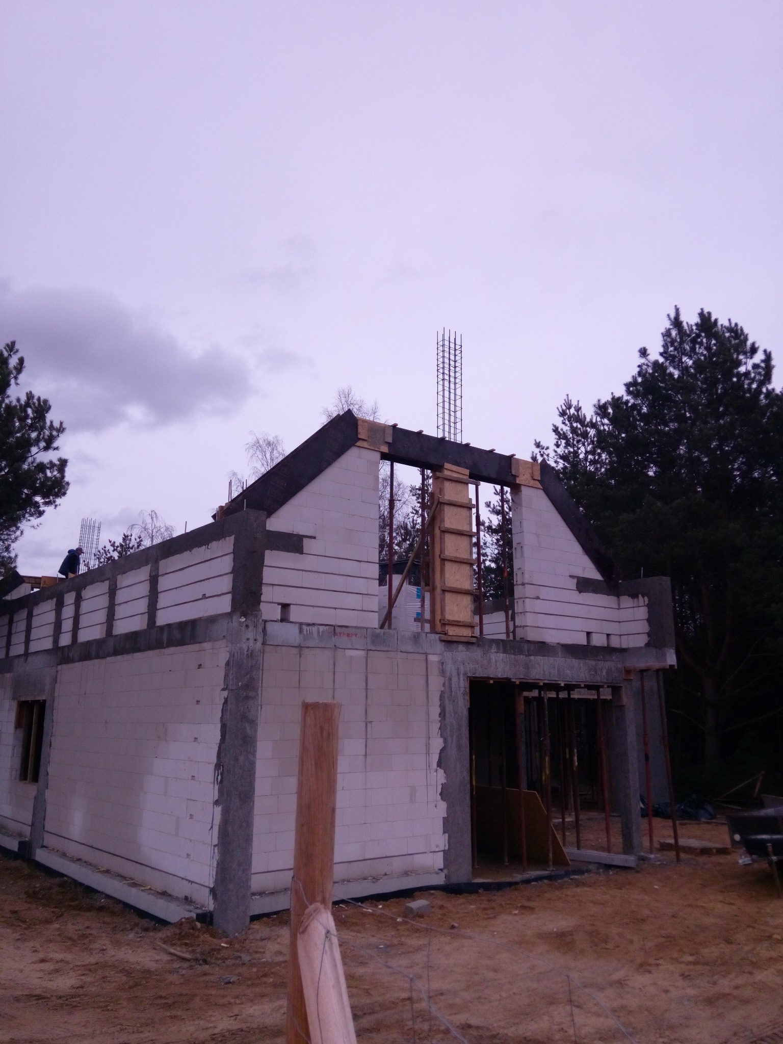 Surowy stan budynku w trakcie budowy: ściany z betonu komórkowego, widoczne zbrojenia pionowe, drewniane szalunki okienne, pochmurne niebo w tle.