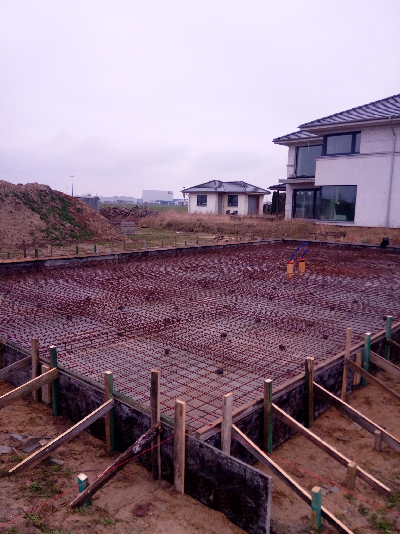 Przygotowany szalunek i zbrojenie płyty fundamentowej pod budowę domu jednorodzinnego, widoczne pionowe rury instalacyjne, w tle hałda ziemi i budynki.