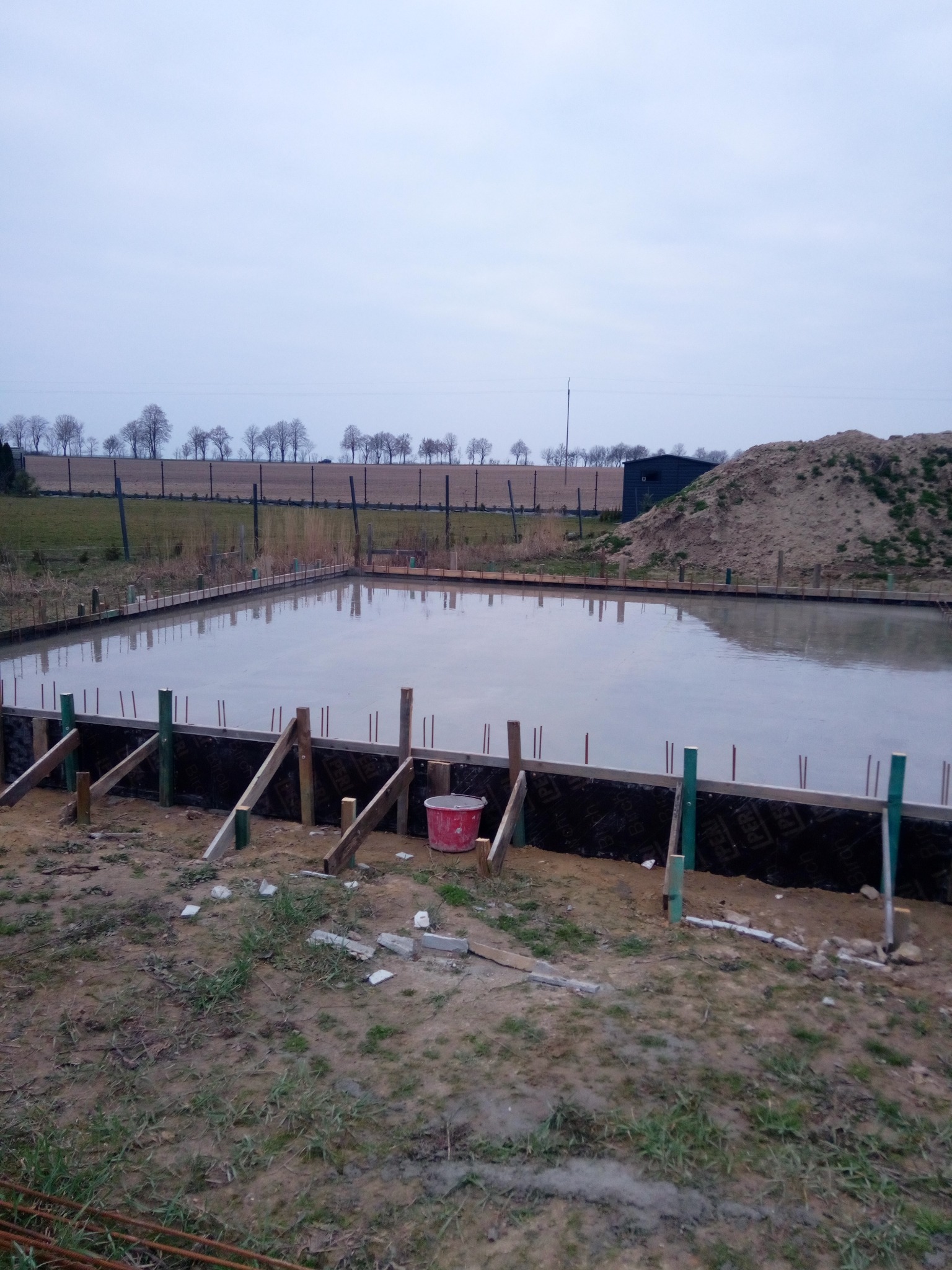 Wylany beton w szalunku pod przyszły budynek, widoczne drewniane podpory, w tle sterta ziemi i budynek gospodarczy.