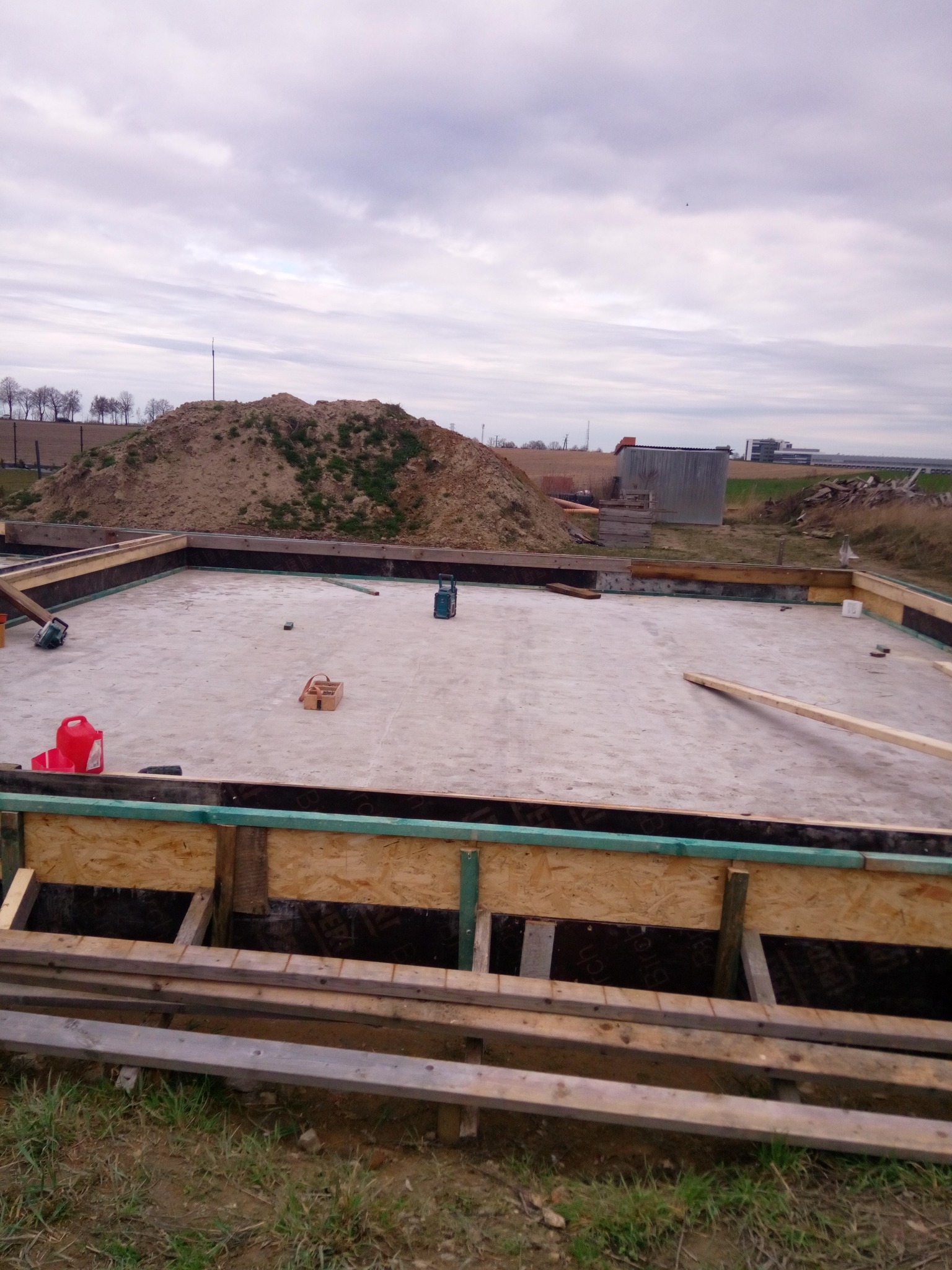 Przygotowany wykop pod wylewkę fundamentową z deskowaniem, widoczny stos drewna i narzędzia, w tle hałda ziemi.