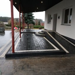 Mar-Bruk - Ułożona ciemnoszara kostka brukowa z białymi akcentami tworząca podjazd i ścieżkę przed domem, widoczny pracownik w odblaskowej kamizelce i zapas kostki w tle.