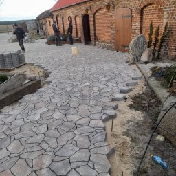 Mar-Bruk - Fragment układanej ścieżki z nieregularnej kostki w odcieniach szarości, z widocznymi pracownikami i stosem kostki w tle, na tle ceglanego budynku z drewnianymi drzwiami.