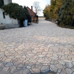 Mar-Bruk - Szeroki, nieregularny chodnik z kostki o nieregularnych kształtach prowadzący w kierunku pola, z budynkiem z czerwonej cegły i drzewami po bokach, w tle widoczne pole.