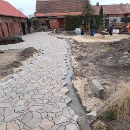 Mar-Bruk - W Pleszewie powstaje ścieżka z nieregularnej kostki, widoczne etapy układania i przygotowywania podłoża, w tle budynki gospodarcze i pracownik z łopatą.