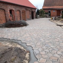 Mar-Bruk - Niedokończona ścieżka z nieregularnych płyt kamiennych w odcieniach beżu i szarości, z widocznym obrzeżem z betonu, na tle czerwonej ceglanej zabudowy gospodarczej z drewnianymi drzwiami.