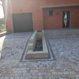 Ułożona kostka brukowa w odcieniach szarości i beżu prowadząca do domu z garażem, z elementami dekoracyjnymi w postaci obramowania z ciemniejszej kostki i podwyższonej rabaty.