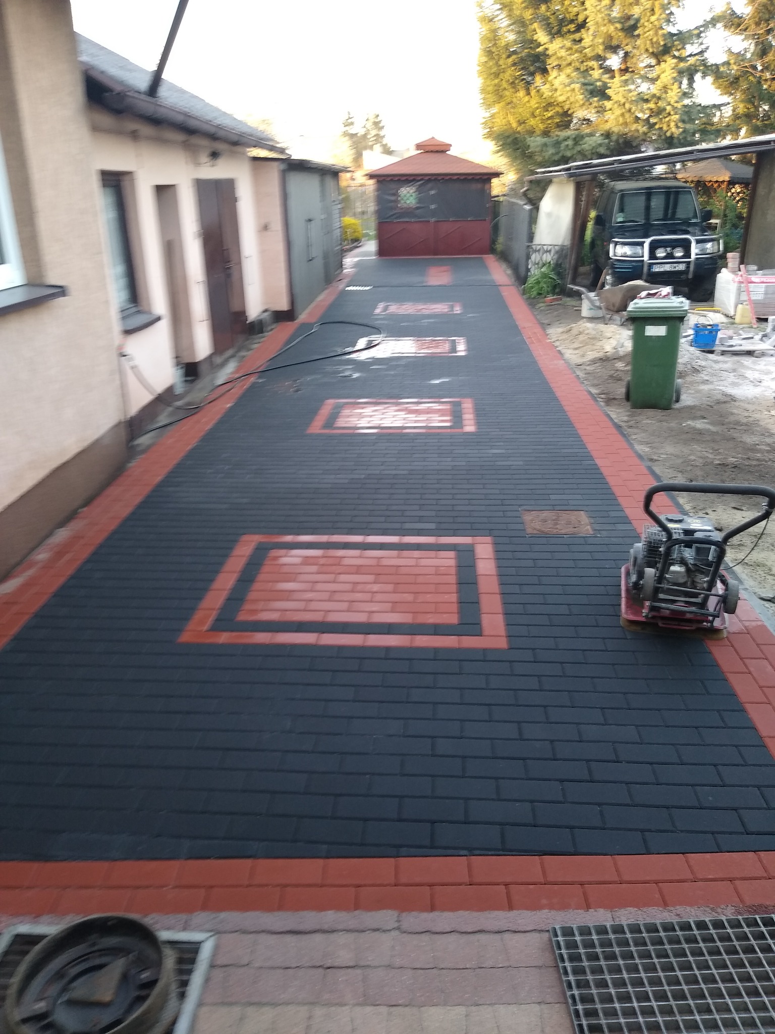 Ułożona czarno-czerwona kostka brukowa z dekoracyjnymi wstawkami w kształcie prostokątów, ubijana zagęszczarką mechaniczną, widoczny fragment domu i podwórza.