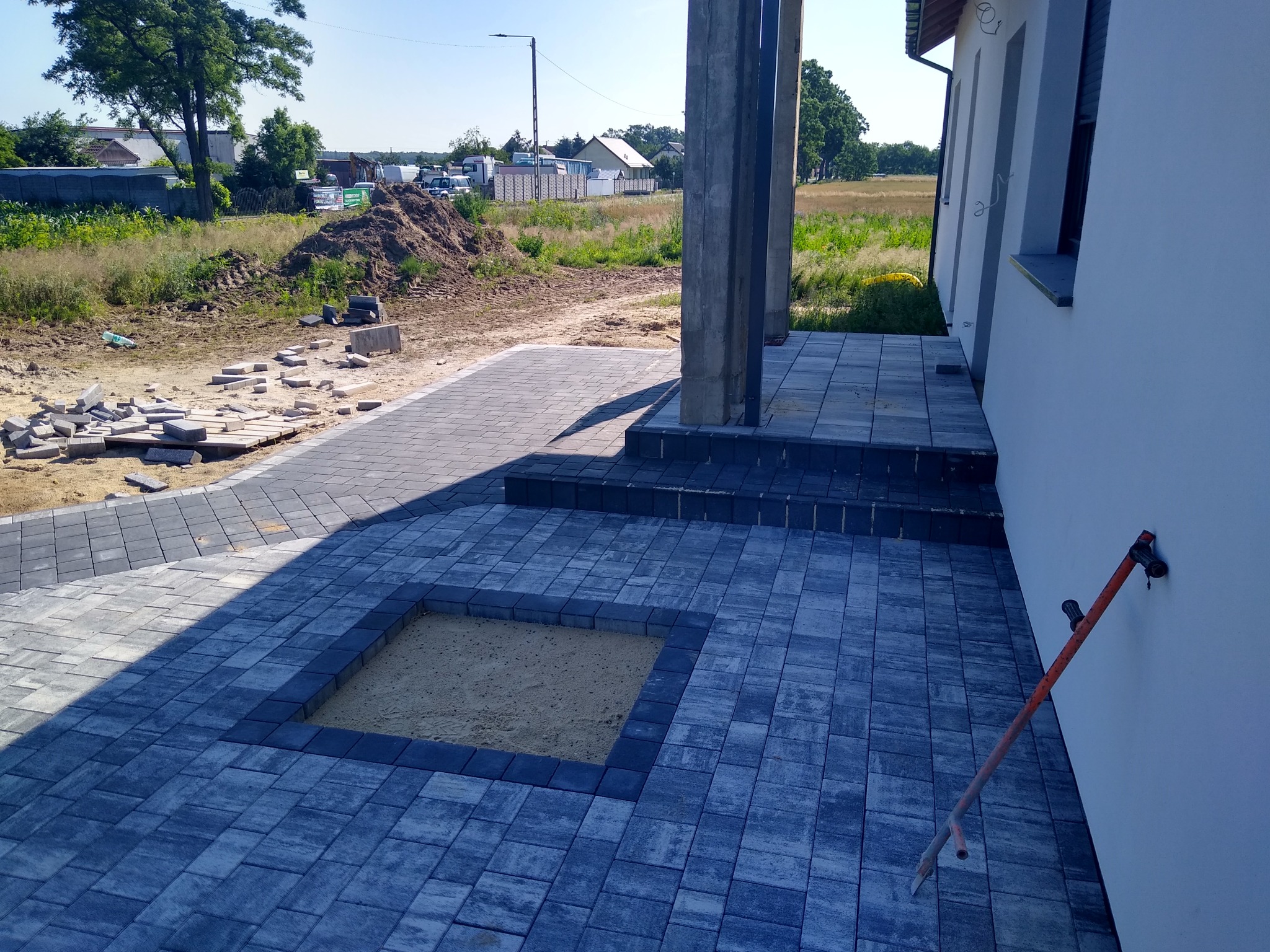 Ułożona, szara kostka brukowa z wyznaczonym miejscem na piaskownicę, widoczna łopata oparta o białą ścianę budynku, w tle przygotowane materiały budowlane.