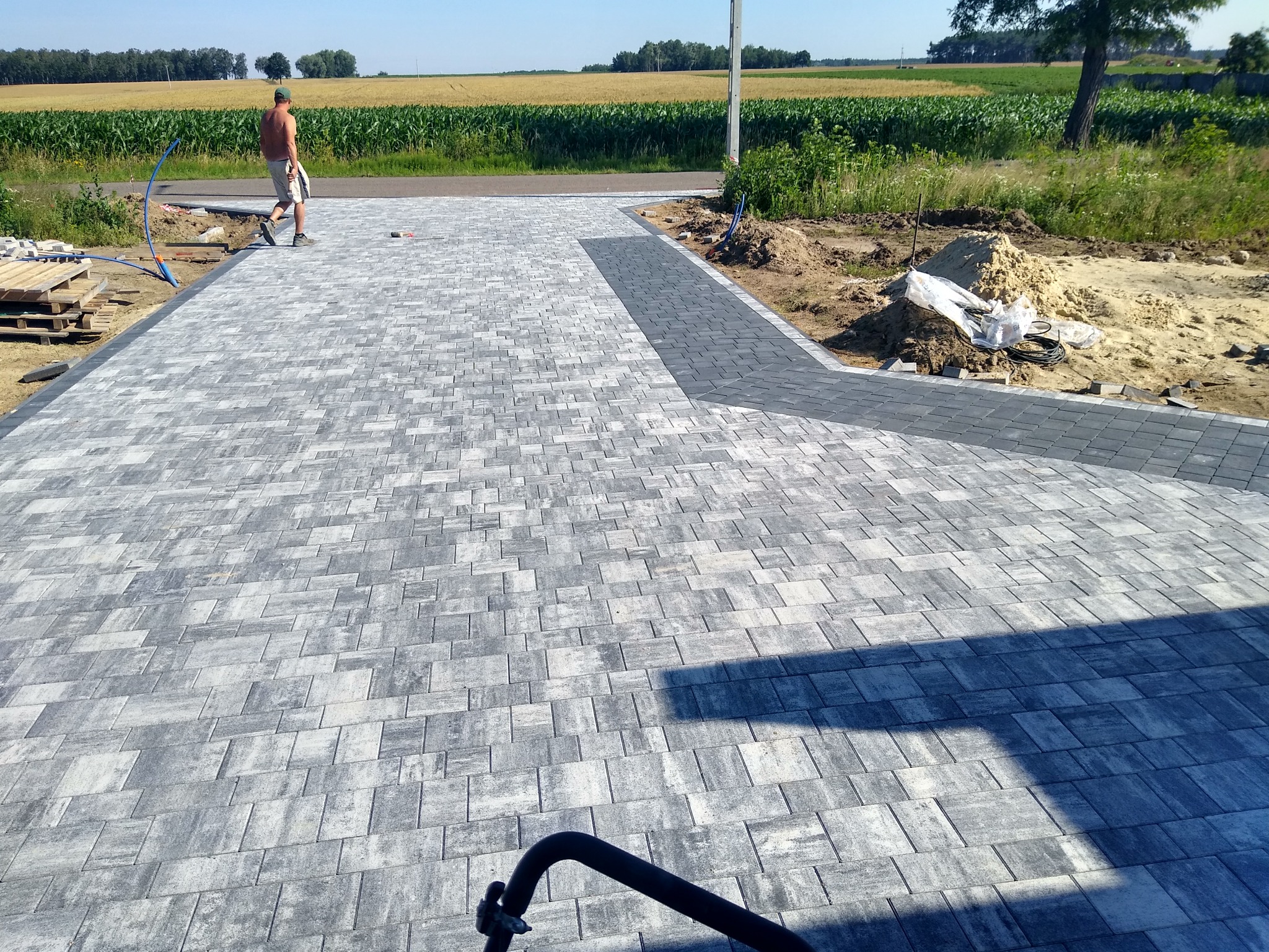 Nowa, szara nawierzchnia z kostki brukowej ułożona na podjeździe, z widocznym pracownikiem w tle oraz stertą piasku i ułożonymi paletami z kostką w pobliżu.