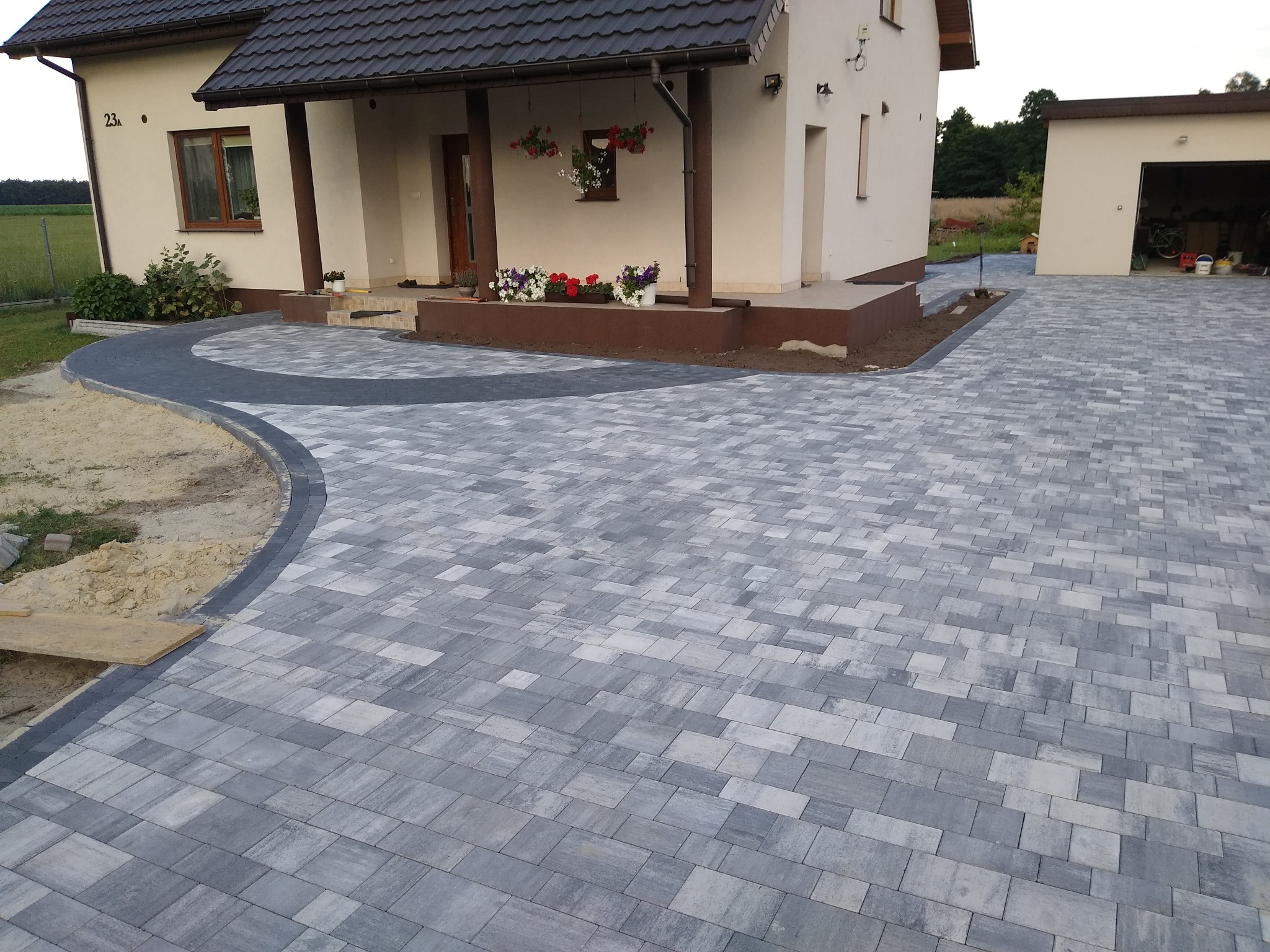 Szara kostka brukowa ułożona na podjeździe przed domem z elementami czarnej kostki oddzielającymi trawnik, widoczny fragment budynku mieszkalnego z kwiatami w donicach na ganku oraz garaż w tle.
