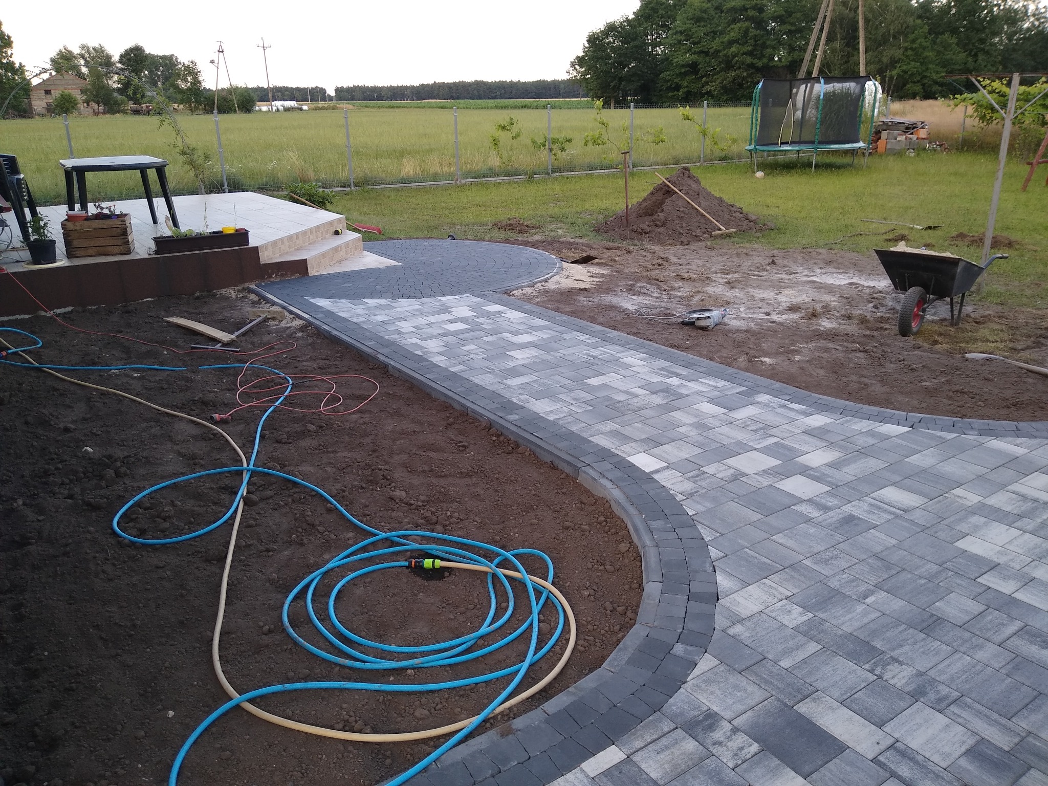 Świeżo ułożona, szara kostka brukowa w kształcie ścieżki i okręgu, z niebieskim wężem ogrodowym na pierwszym planie i widokiem na zielone pole w tle.