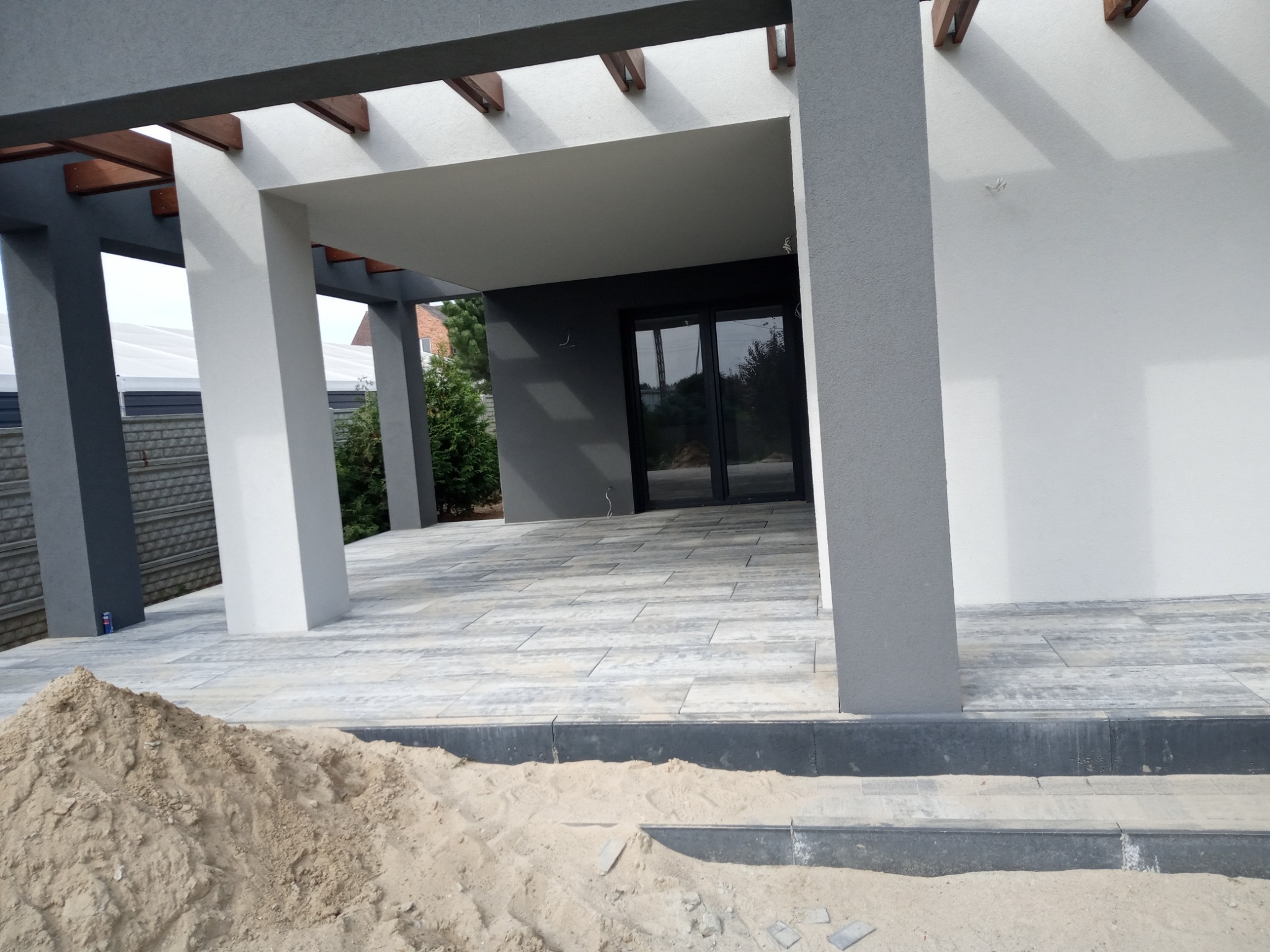 Nowoczesny taras z kostki brukowej w odcieniach szarości, z widocznym stosem piasku potrzebnego do prac wykończeniowych.