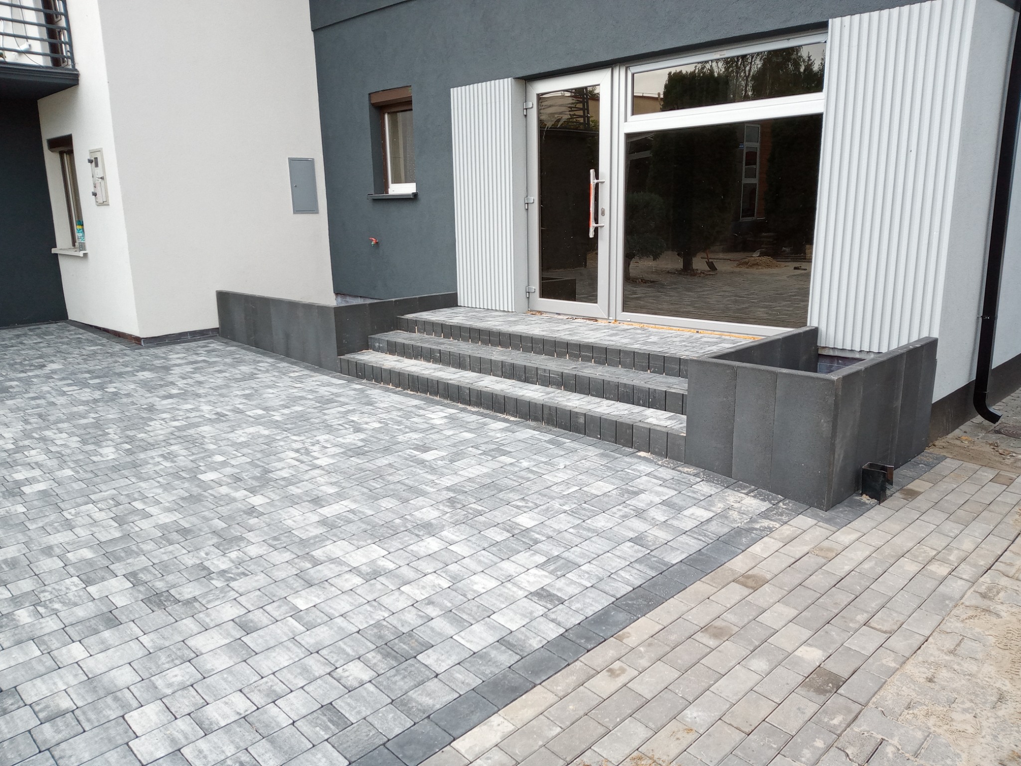Ułożona szara kostka brukowa przed nowoczesnym budynkiem z szarymi ścianami i przeszklonymi drzwiami wejściowymi, widoczne schody z kostki brukowej i fragmenty kostki granitowej.