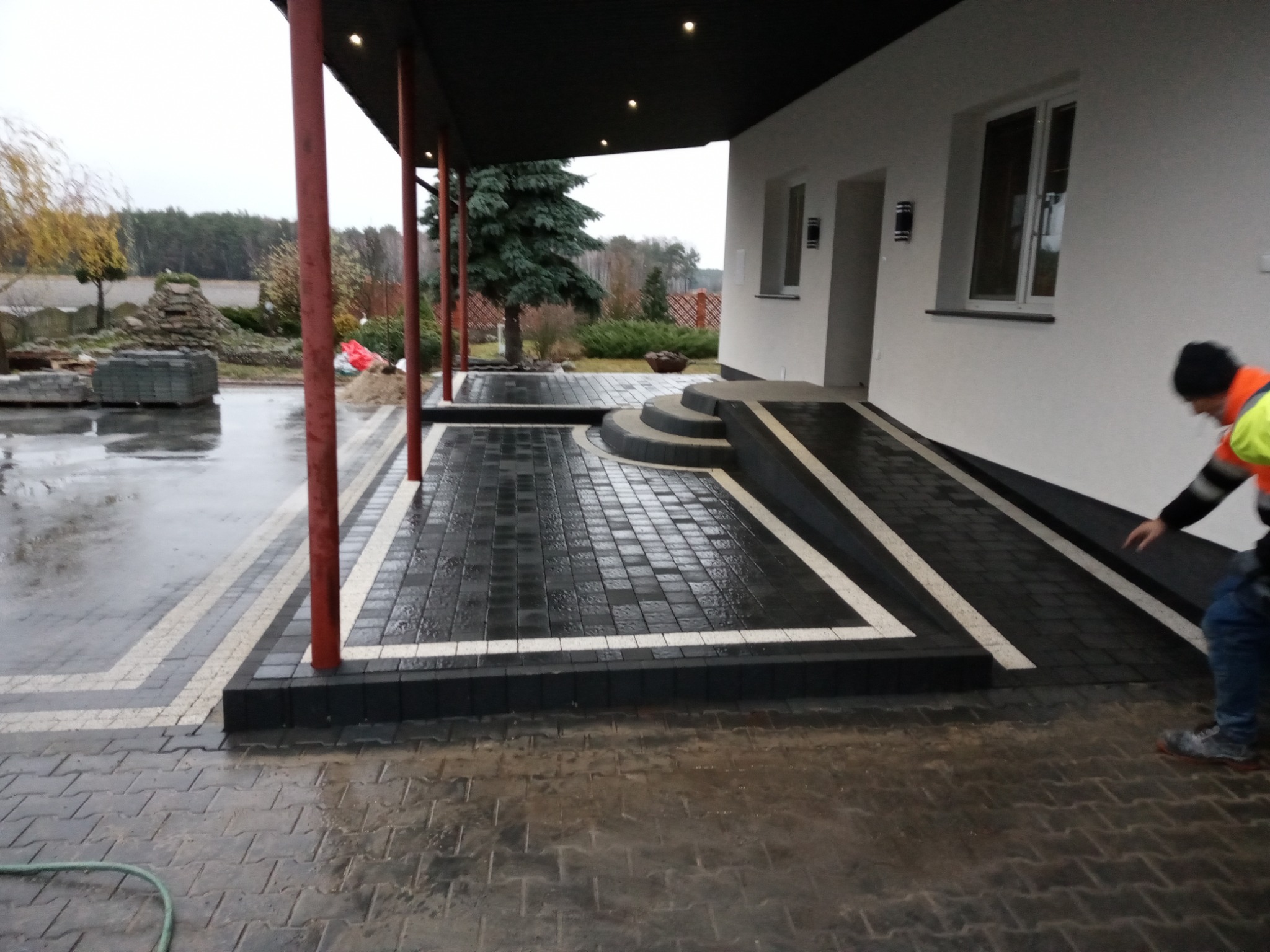 Ułożona ciemnoszara kostka brukowa z białymi akcentami tworząca podjazd i ścieżkę przed domem, widoczny pracownik w odblaskowej kamizelce i zapas kostki w tle.