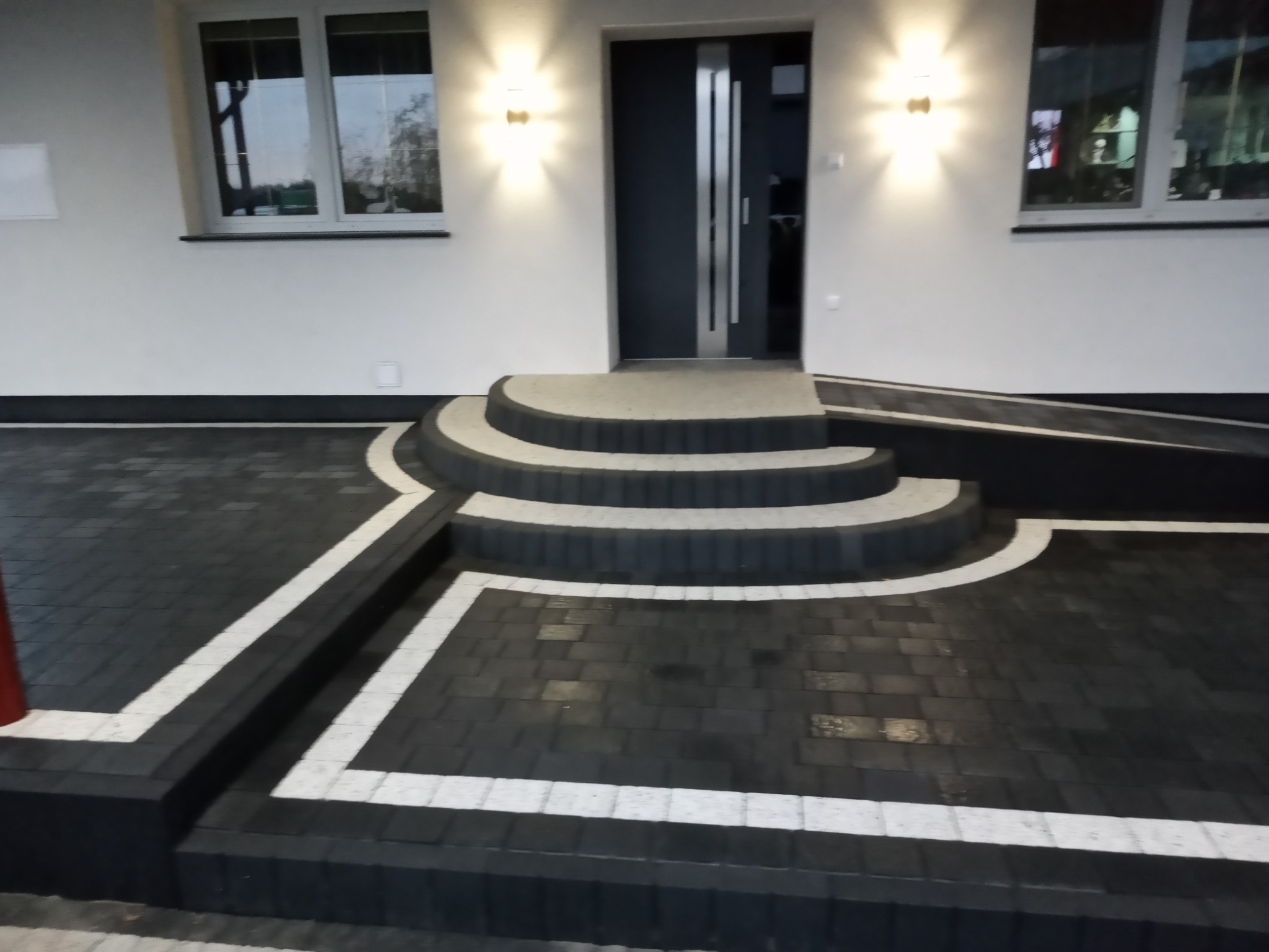 Nowoczesne wejście do domu z ciemnej kostki brukowej z białymi obwódkami, schody półokrągłe prowadzące do drzwi wejściowych, oświetlone lampami po bokach.