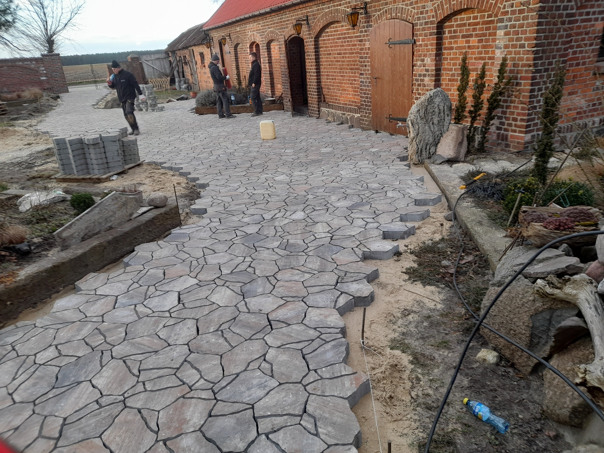 Fragment układanej ścieżki z nieregularnej kostki w odcieniach szarości, z widocznymi pracownikami i stosem kostki w tle, na tle ceglanego budynku z drewnianymi drzwiami.
