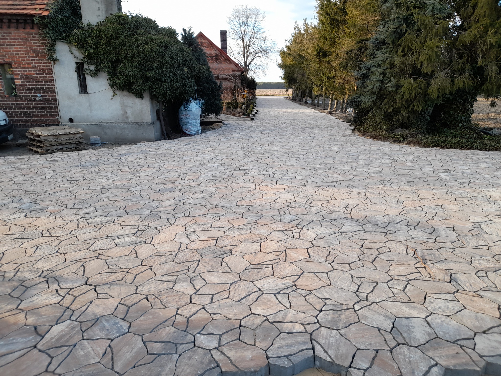 Szeroki, nieregularny chodnik z kostki o nieregularnych kształtach prowadzący w kierunku pola, z budynkiem z czerwonej cegły i drzewami po bokach, w tle widoczne pole.