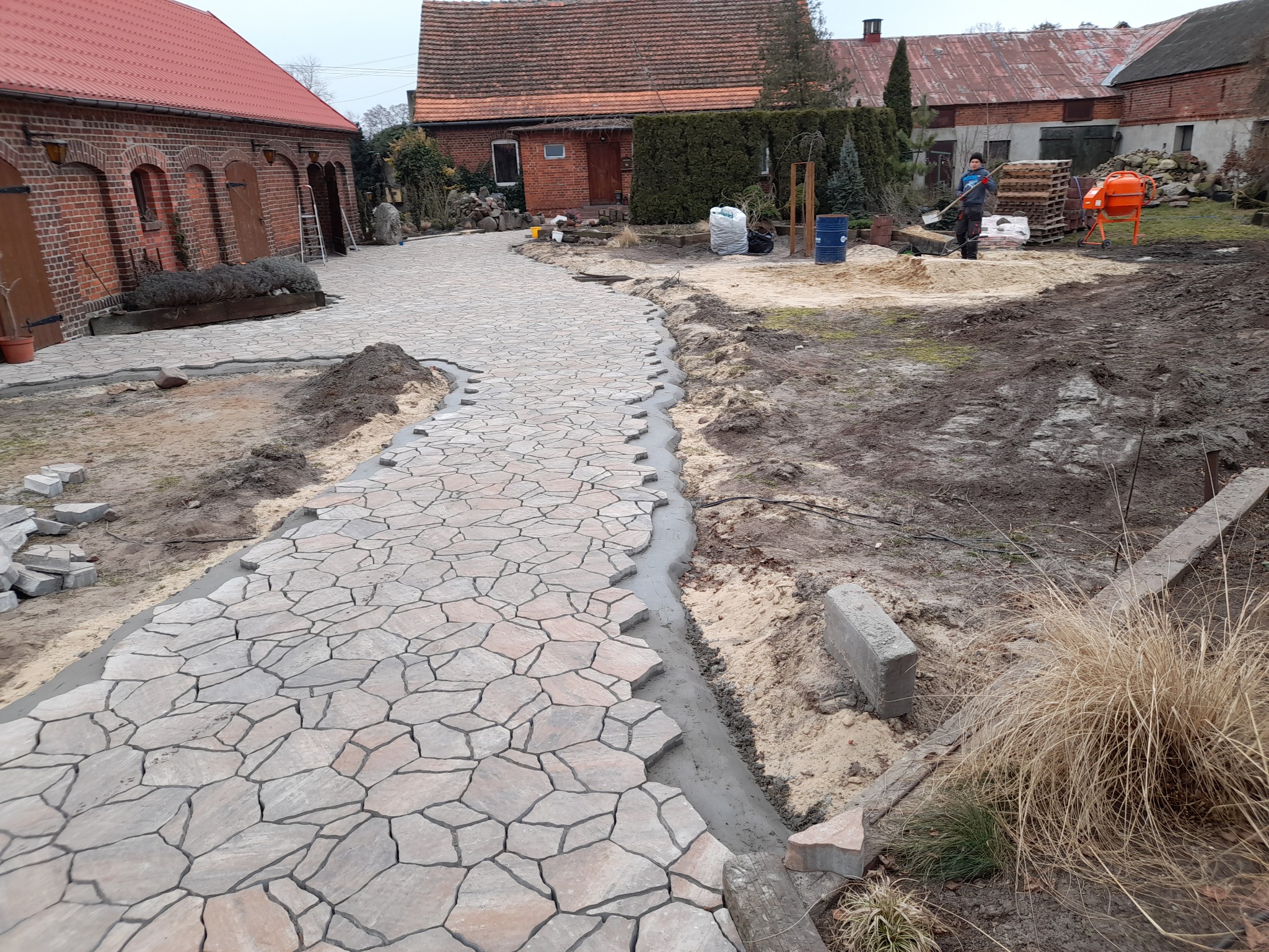 W Pleszewie powstaje ścieżka z nieregularnej kostki, widoczne etapy układania i przygotowywania podłoża, w tle budynki gospodarcze i pracownik z łopatą.