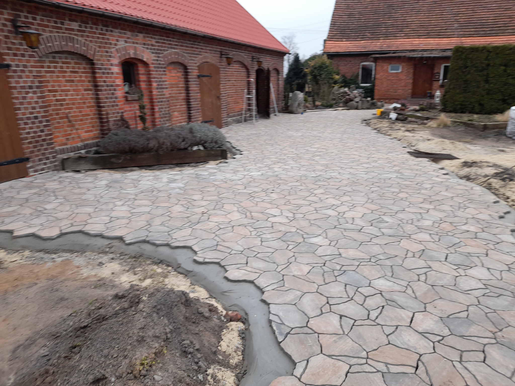 Niedokończona ścieżka z nieregularnych płyt kamiennych w odcieniach beżu i szarości, z widocznym obrzeżem z betonu, na tle czerwonej ceglanej zabudowy gospodarczej z drewnianymi drzwiami.