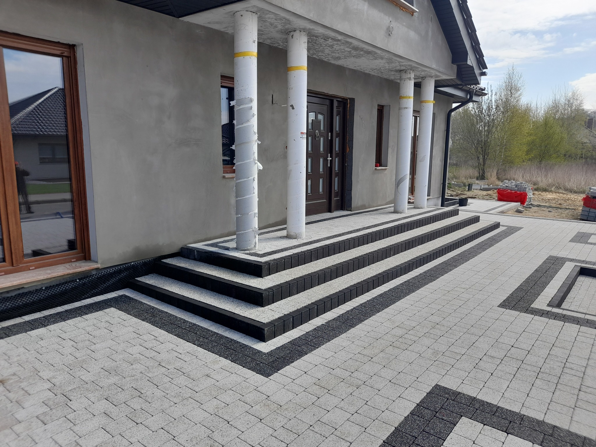 Nowo ułożona, dwukolorowa kostka brukowa przed domem z kolumnami, schodami i widocznymi materiałami budowlanymi w tle.