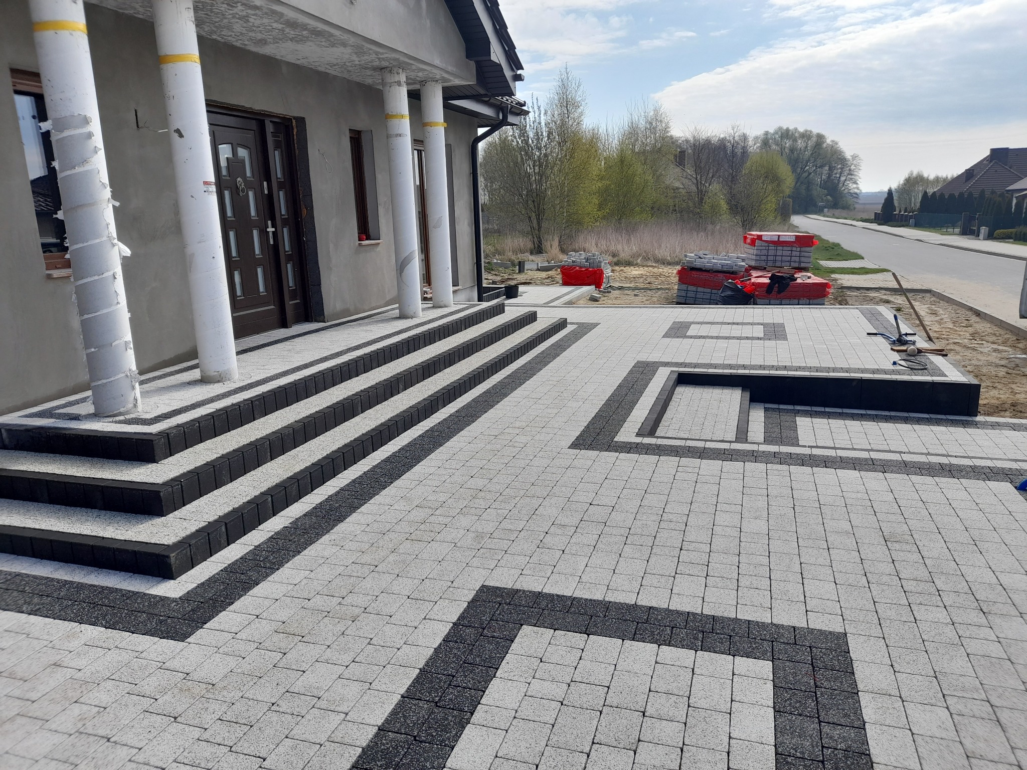 Ułożona kostka brukowa w dwóch kolorach (szary i grafitowy) tworząca wzór przed domem z kolumnami. Na drugim planie sterta kostki brukowej i ulica.