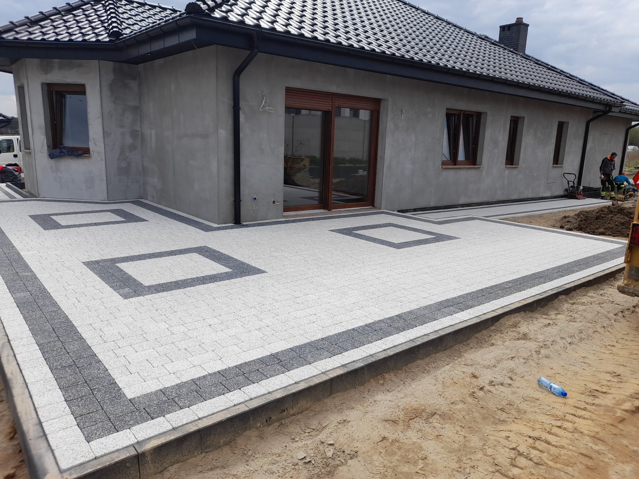 Nowo ułożony chodnik z kostki brukowej w geometryczny wzór przed szarym domem z ciemną dachówką, widoczny pracownik budowlany z ubijarką w tle.