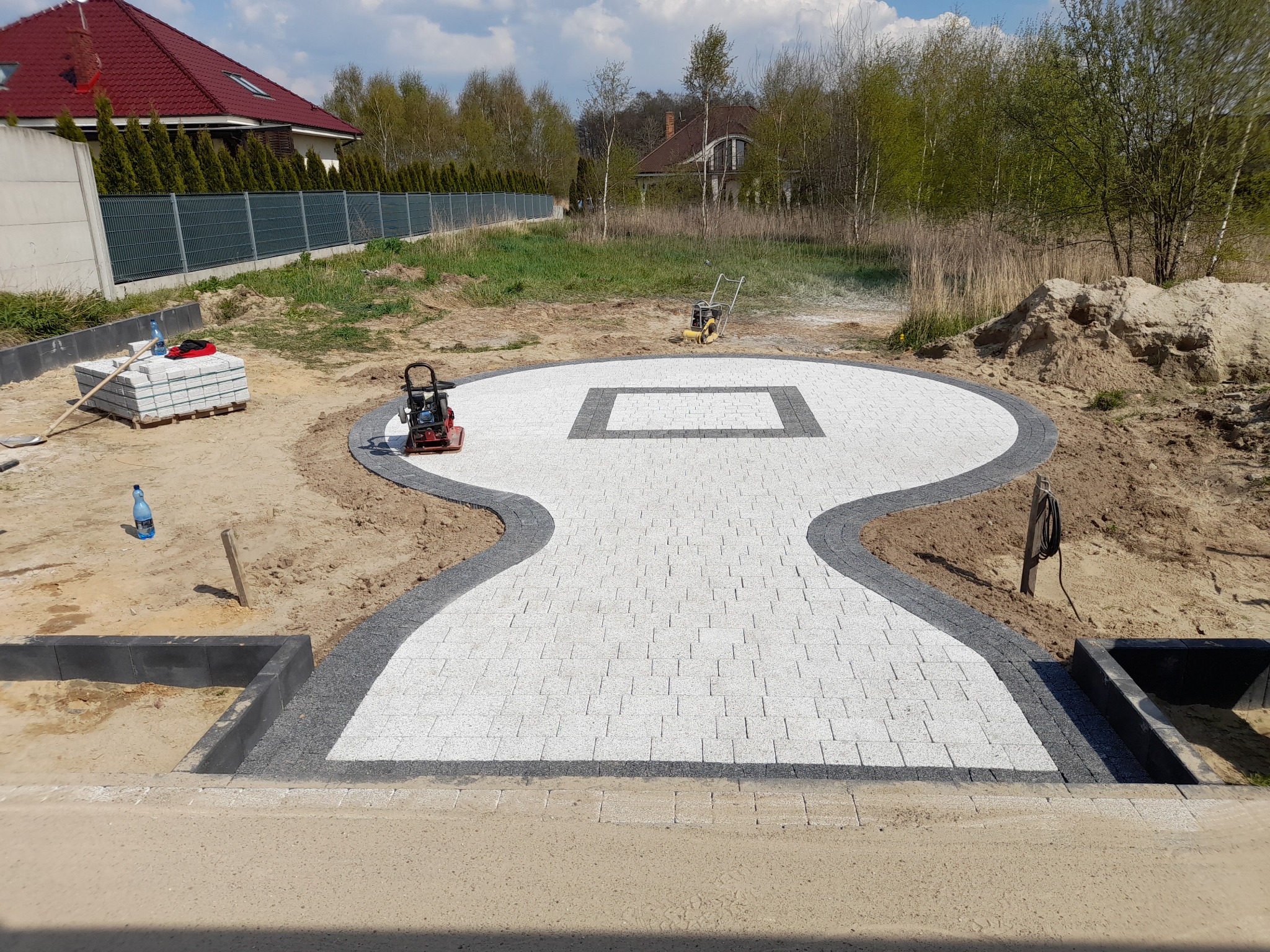 Kształtna ścieżka z jasnoszarej kostki brukowej z ciemnoszarą obwódką w trakcie układania, widoczne maszyny do ubijania ziemi, paleta kostki i butelka wody mineralnej.